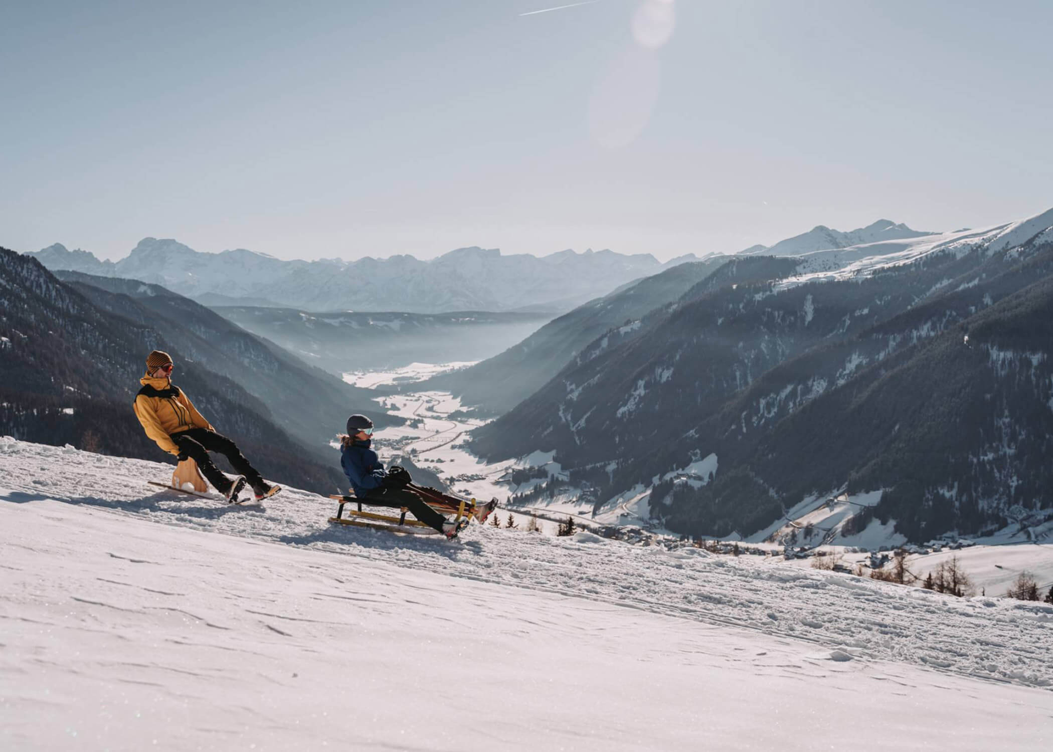 Zwei Personen fahren mit dem Schlitten einen verschneiten Berghang hinunter, mit einem malerischen Tal und Bergen im Hintergrund. - Hotel La Casies Mountain Living