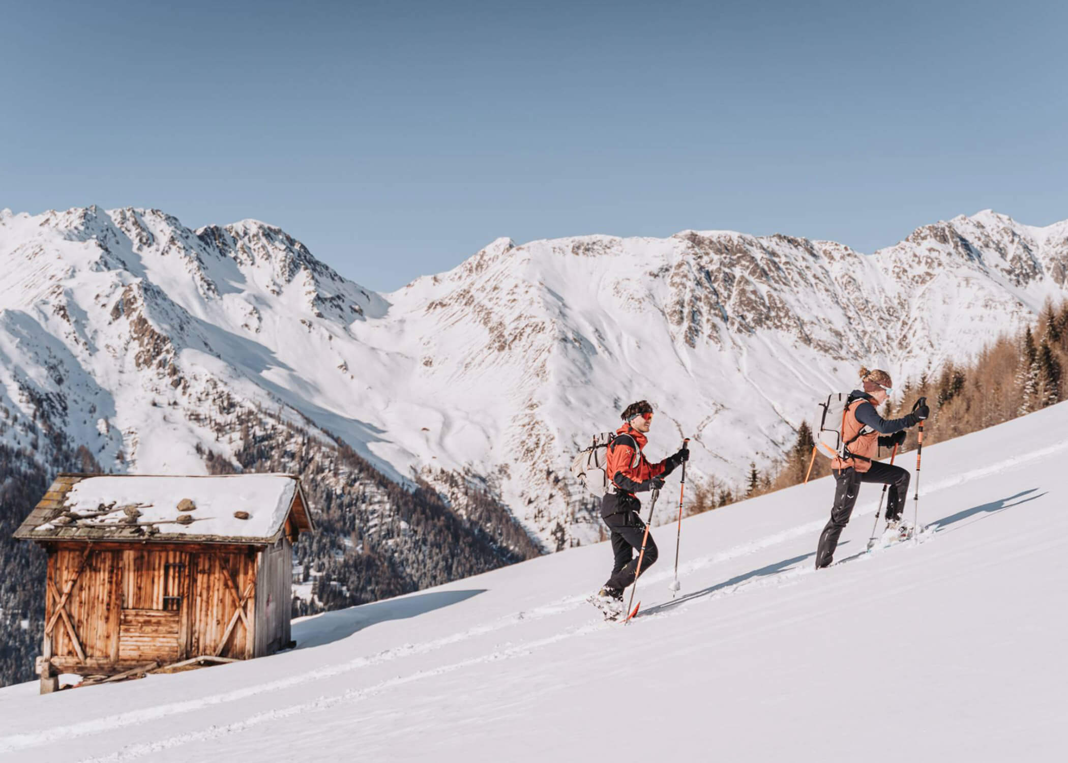 Zwei Personen wandern mit Stöcken bergauf auf Schnee in der Nähe einer Holzhütte, im Hintergrund verschneite Berge. - Hotel La Casies Mountain Living