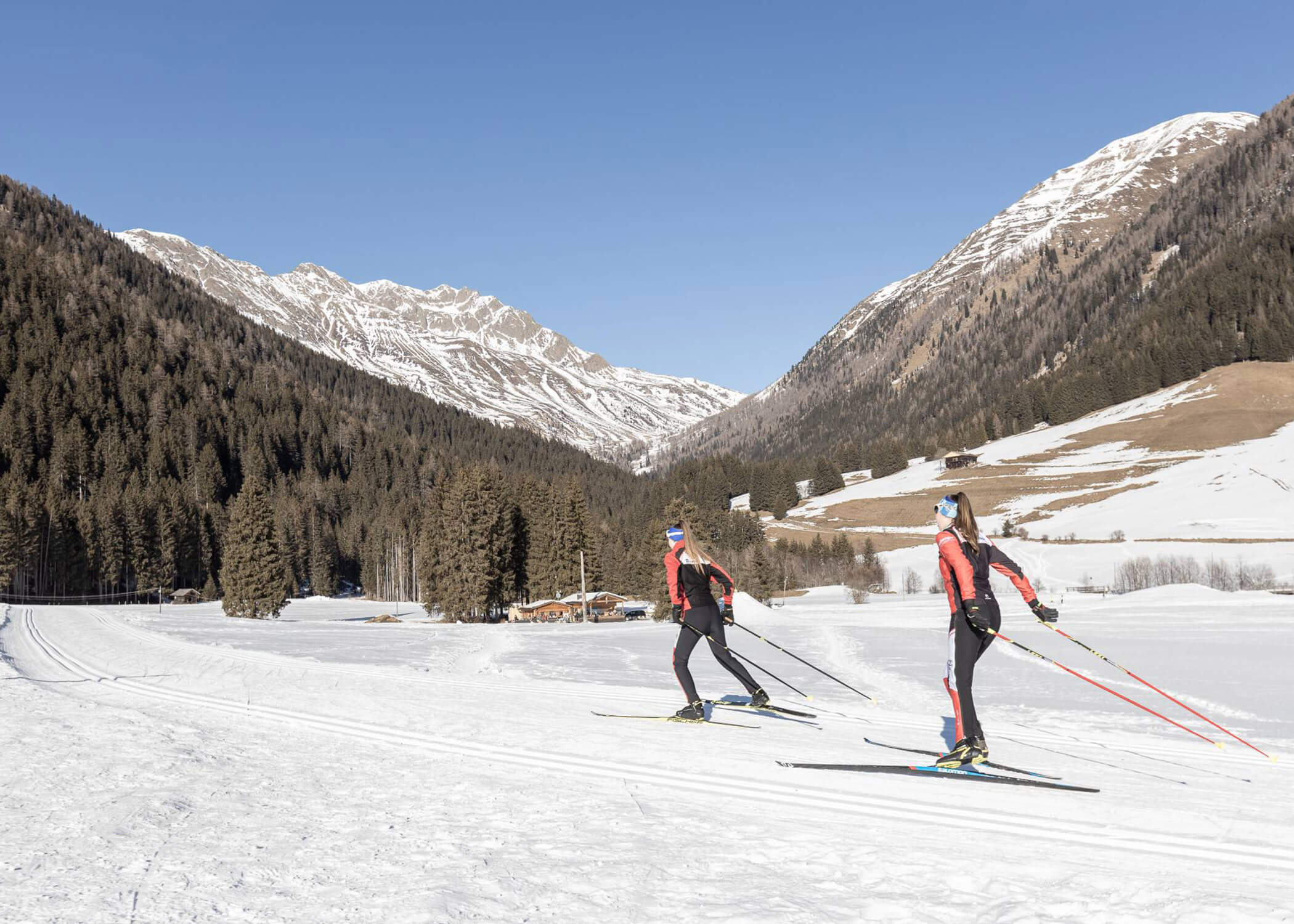Zwei Personen beim Skilanglauf auf einer verschneiten Loipe, umgeben von Bergen und Kiefern unter einem klaren Himmel. - Hotel La Casies Mountain Living
