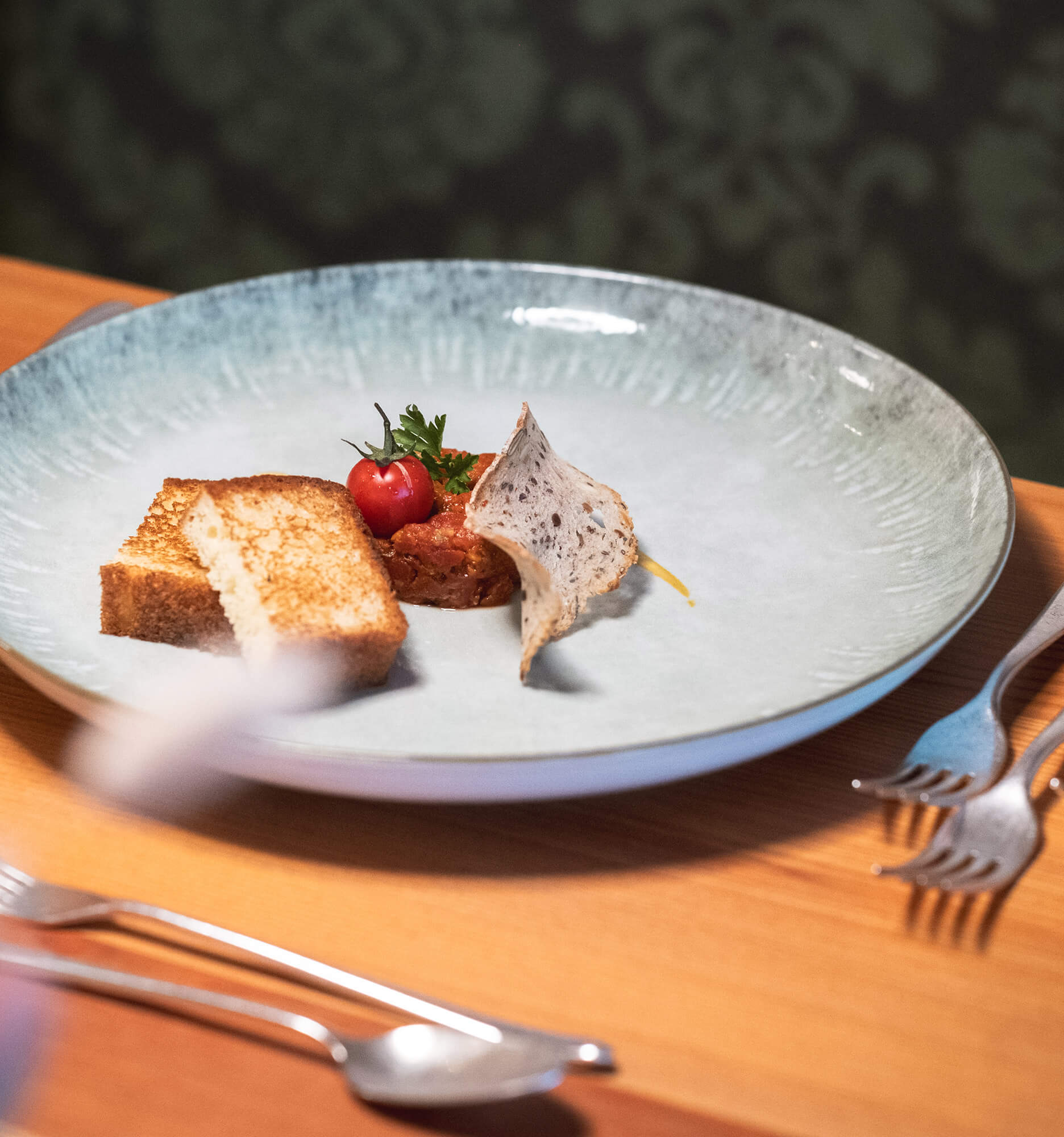 Eine plattierte Gourmet-Vorspeise mit Toast, Tomatenbeilage und einem dünnen Knusperstück auf einem hellblauen Keramikteller. - Hotel La Casies Mountain Living