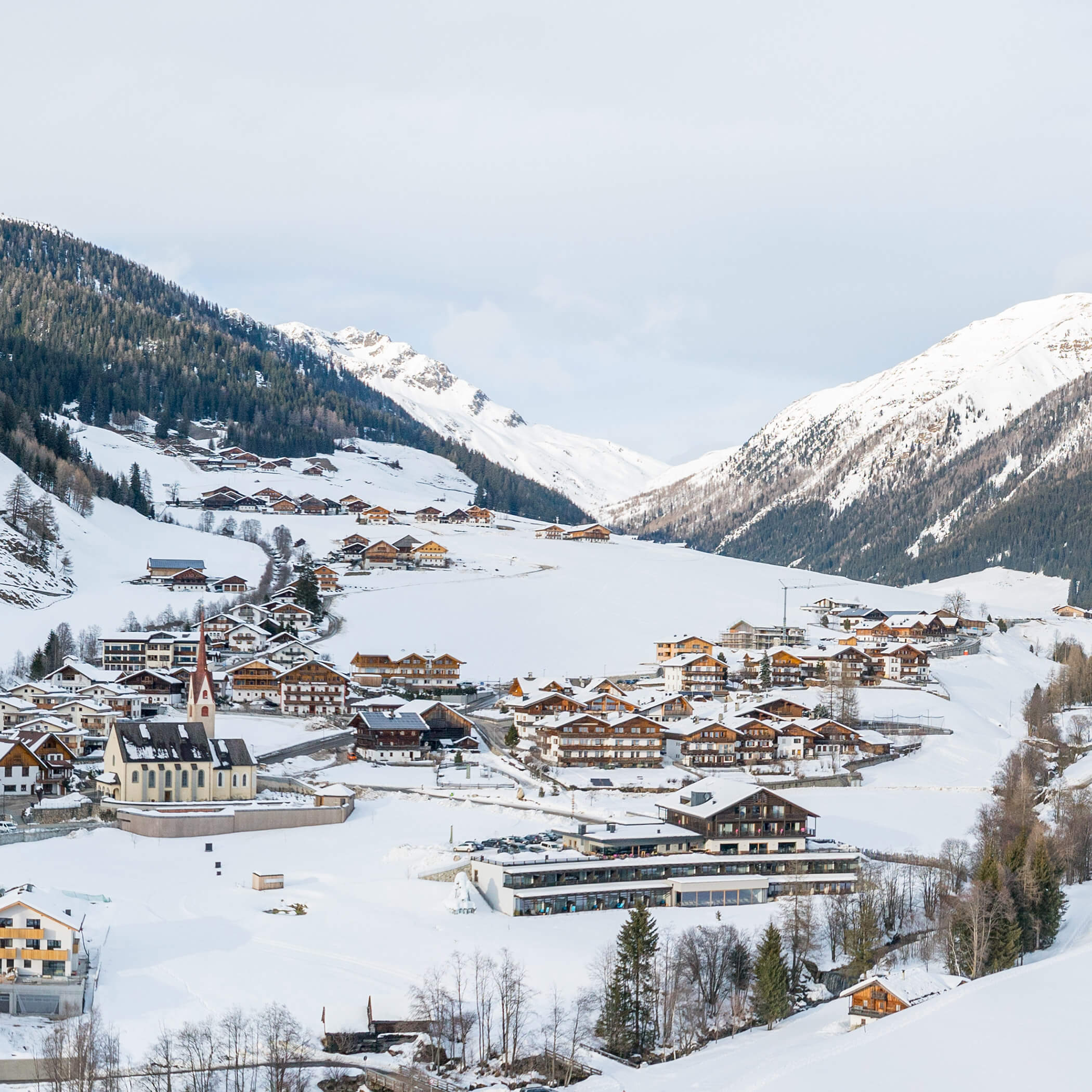 Schneebedecktes Alpendorf mit verstreuten Häusern, einer Kirche und Bergen im Hintergrund. - Hotel La Casies Mountain Living
