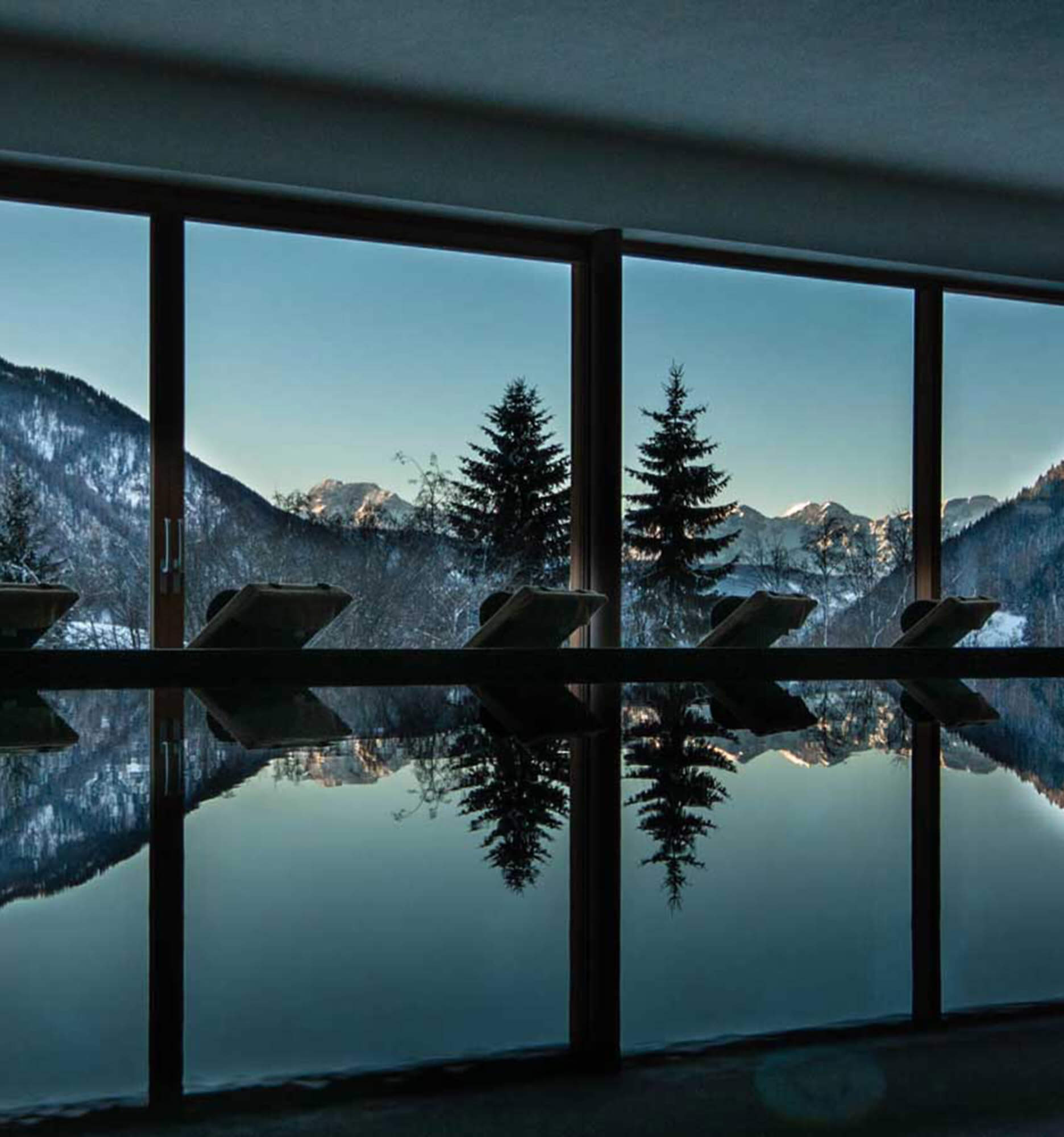 Innenpool mit Liegestühlen, großen Fenstern und einer Berglandschaft mit Kiefern, die sich im Wasser spiegeln. - Hotel La Casies Mountain Living