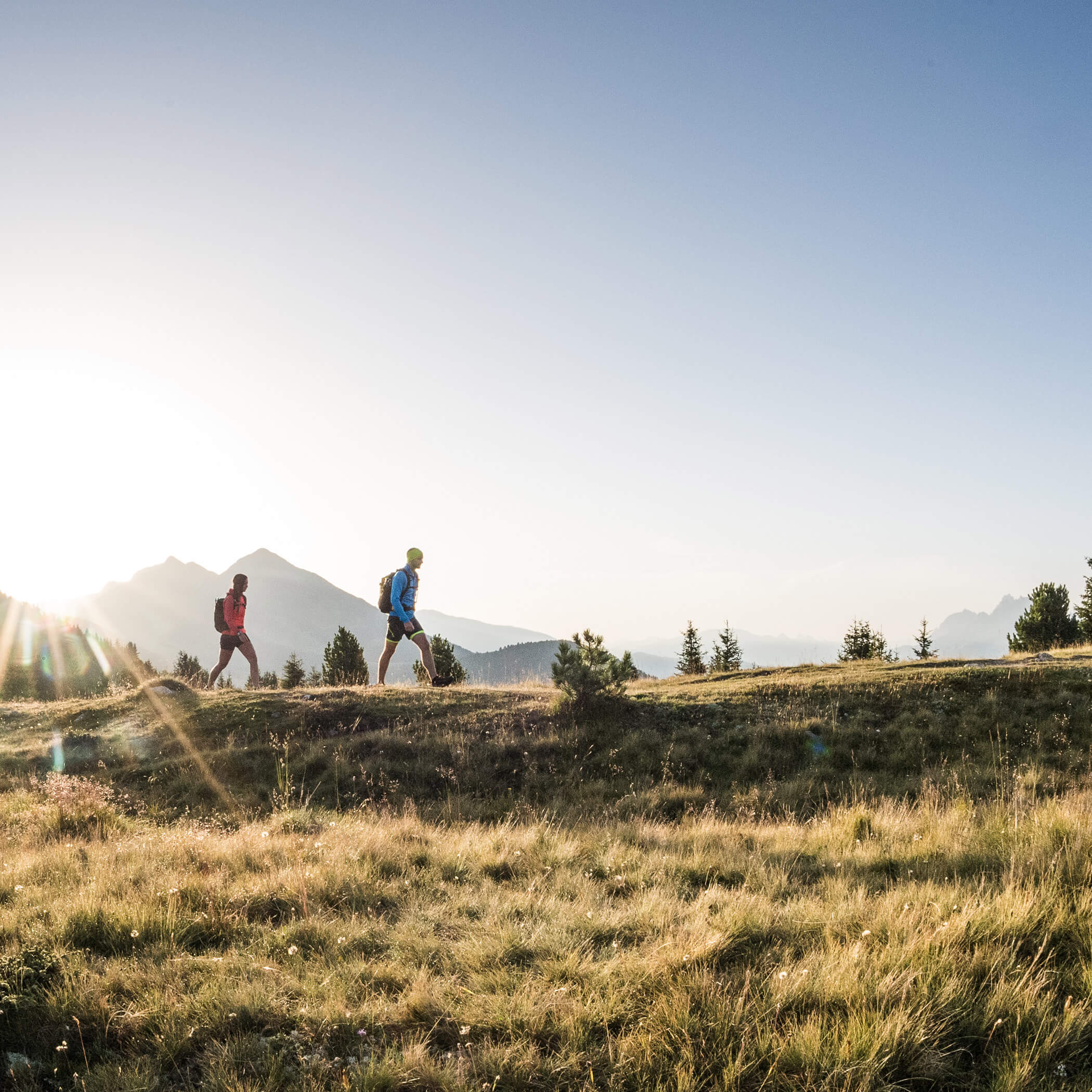 Zwei Wanderer mit Rucksäcken gehen bei Sonnenaufgang über einen grasbewachsenen Weg mit Bergen im Hintergrund. - Hotel La Casies Mountain Living