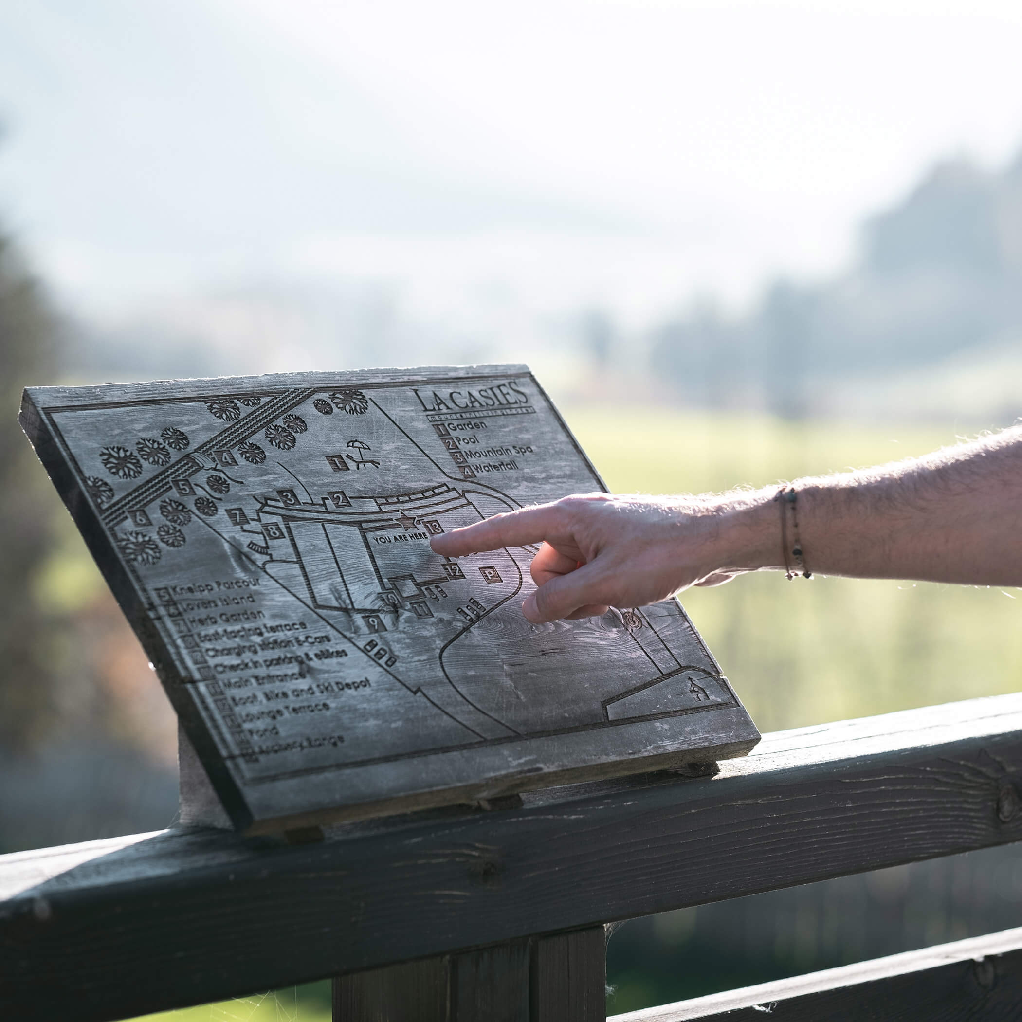 Eine Person zeigt auf eine hölzerne Karte mit der Aufschrift LACASIES an einem Balkongeländer im Sonnenlicht. - Hotel La Casies Mountain Living
