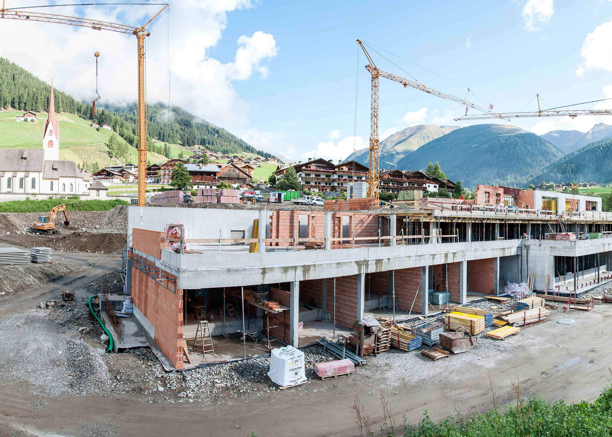 Großes Gebäude im Bau mit Kränen in einem Bergdorf; Kirche und Häuser im Hintergrund. - Hotel La Casies Mountain Living