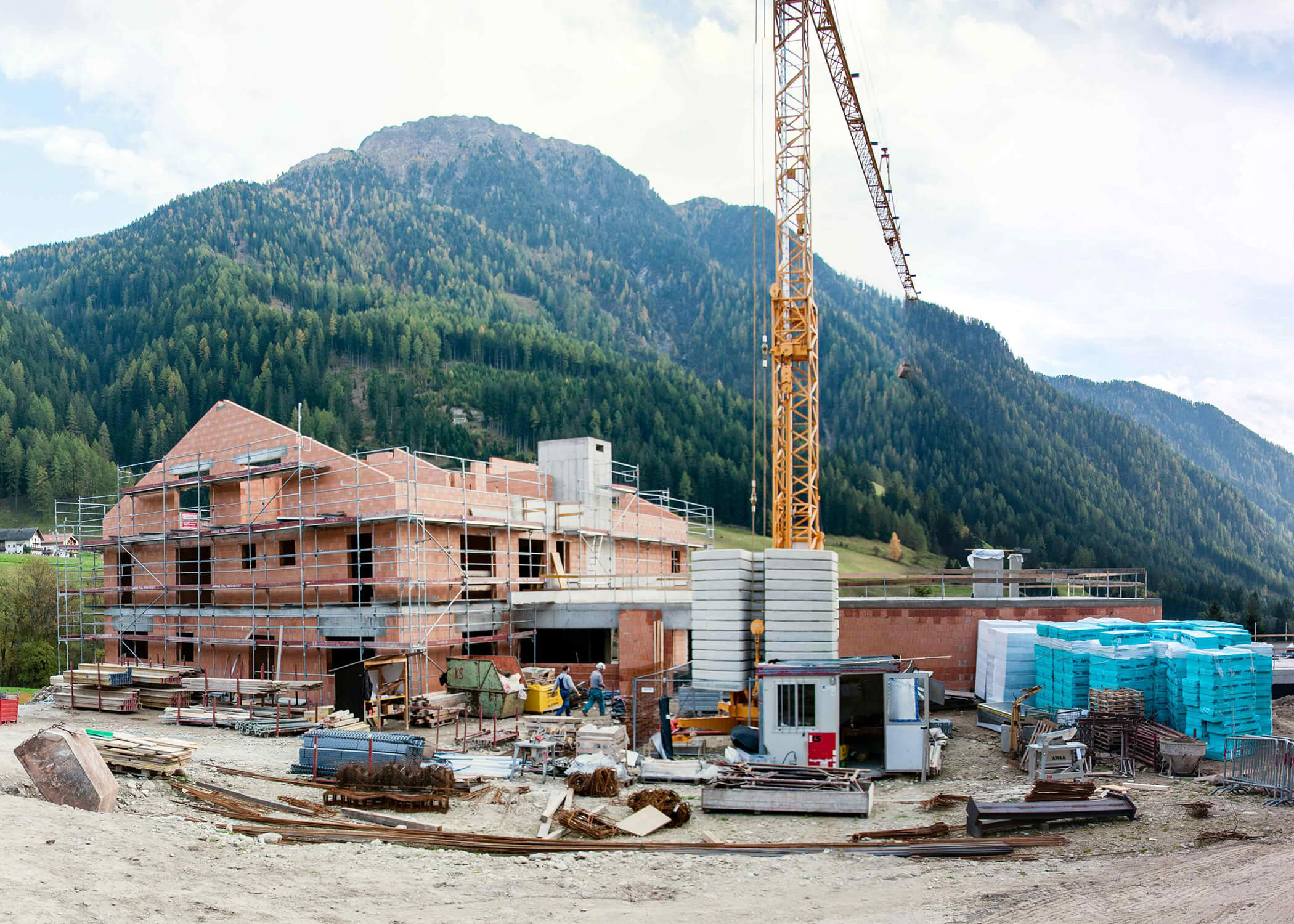 Baustelle mit unfertigem Ziegelgebäude, Kran und baumbewachsenen Bergen im Hintergrund. - Hotel La Casies Mountain Living