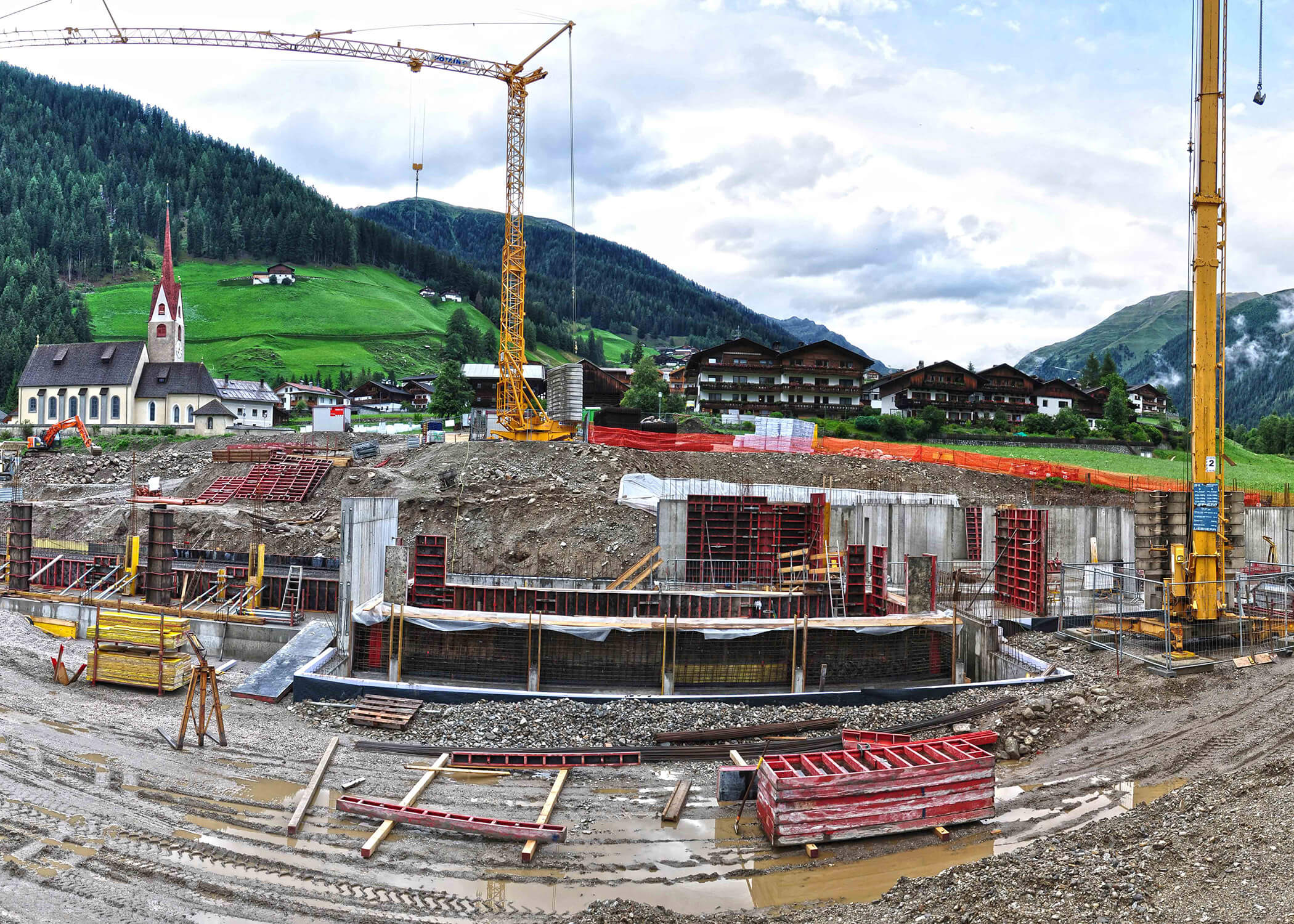 Baustelle mit Kränen und Baumaterialien in einem Bergdorf, Kirche und Häuser im Hintergrund. - Hotel La Casies Mountain Living