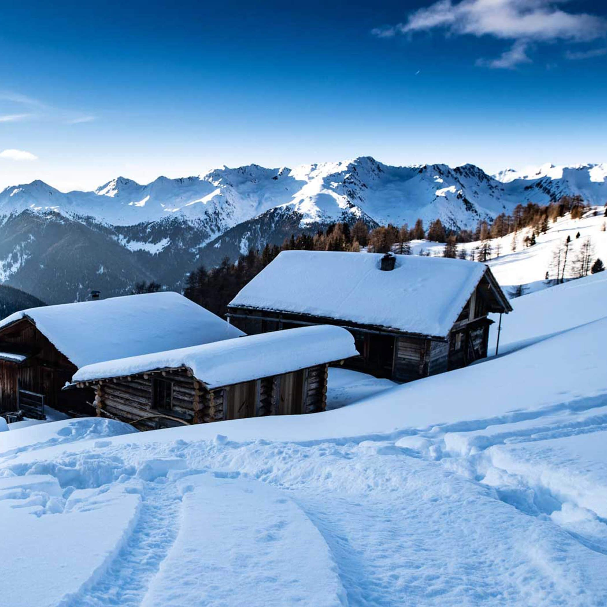 Snow-covered wooden cabins on a mountain slope with snowy peaks and blue sky in the background. - Hotel La Casies Mountain Living