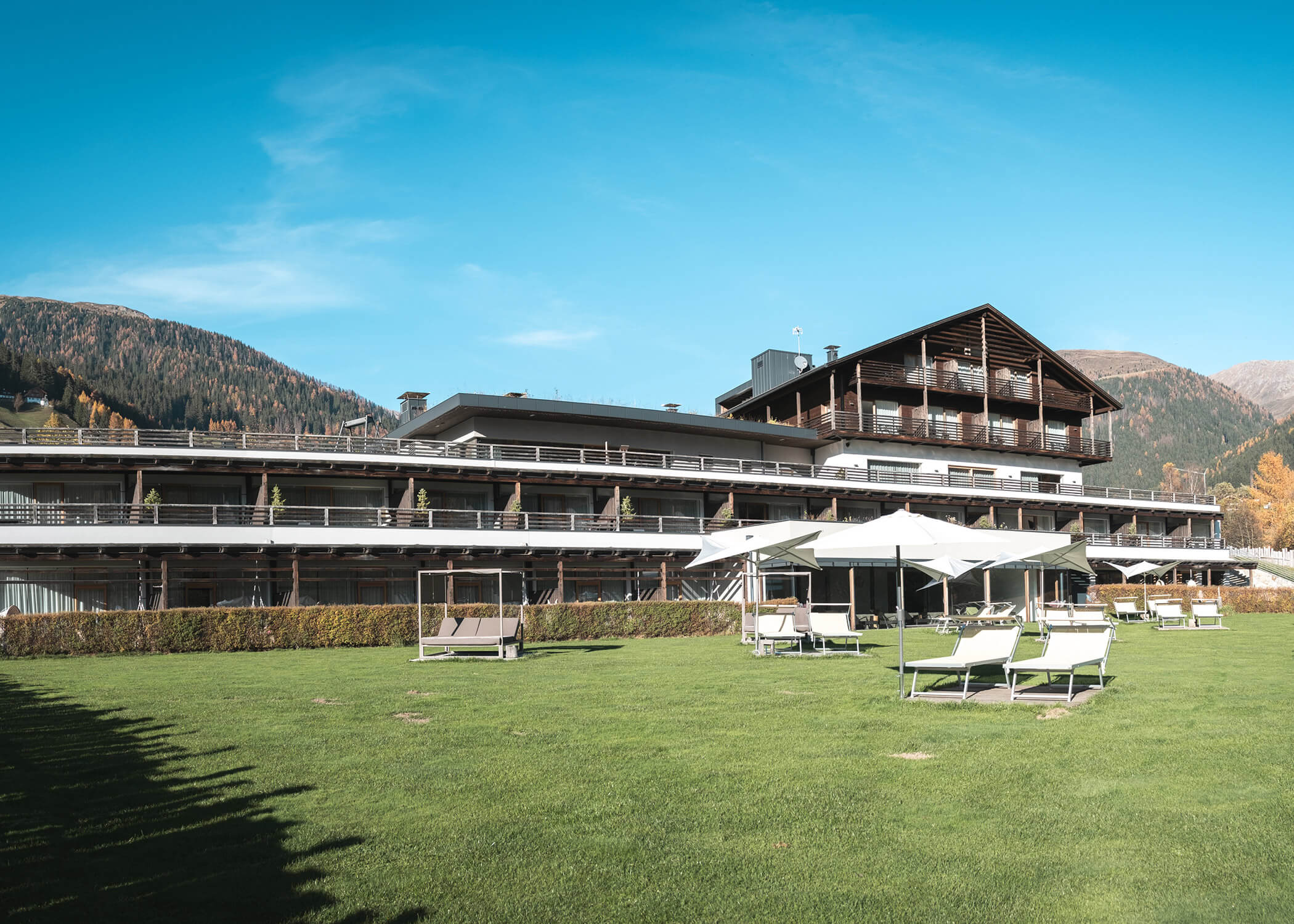 Modernes Hotel mit großen Balkonen und Liegestühlen auf einer grünen Wiese, umgeben von Bergen und blauem Himmel. - Hotel La Casies Mountain Living