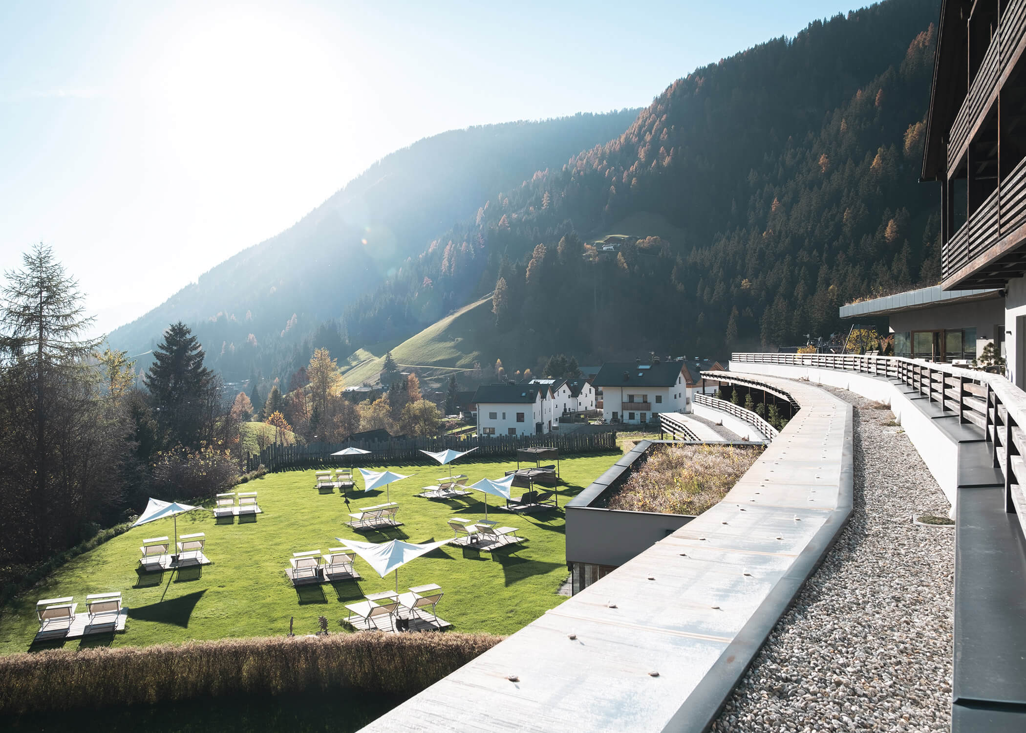 Grüner Rasen mit Liegestühlen und weißen Schirmen vor der Kulisse bewaldeter Berge in hellem Sonnenlicht. - Hotel La Casies Mountain Living