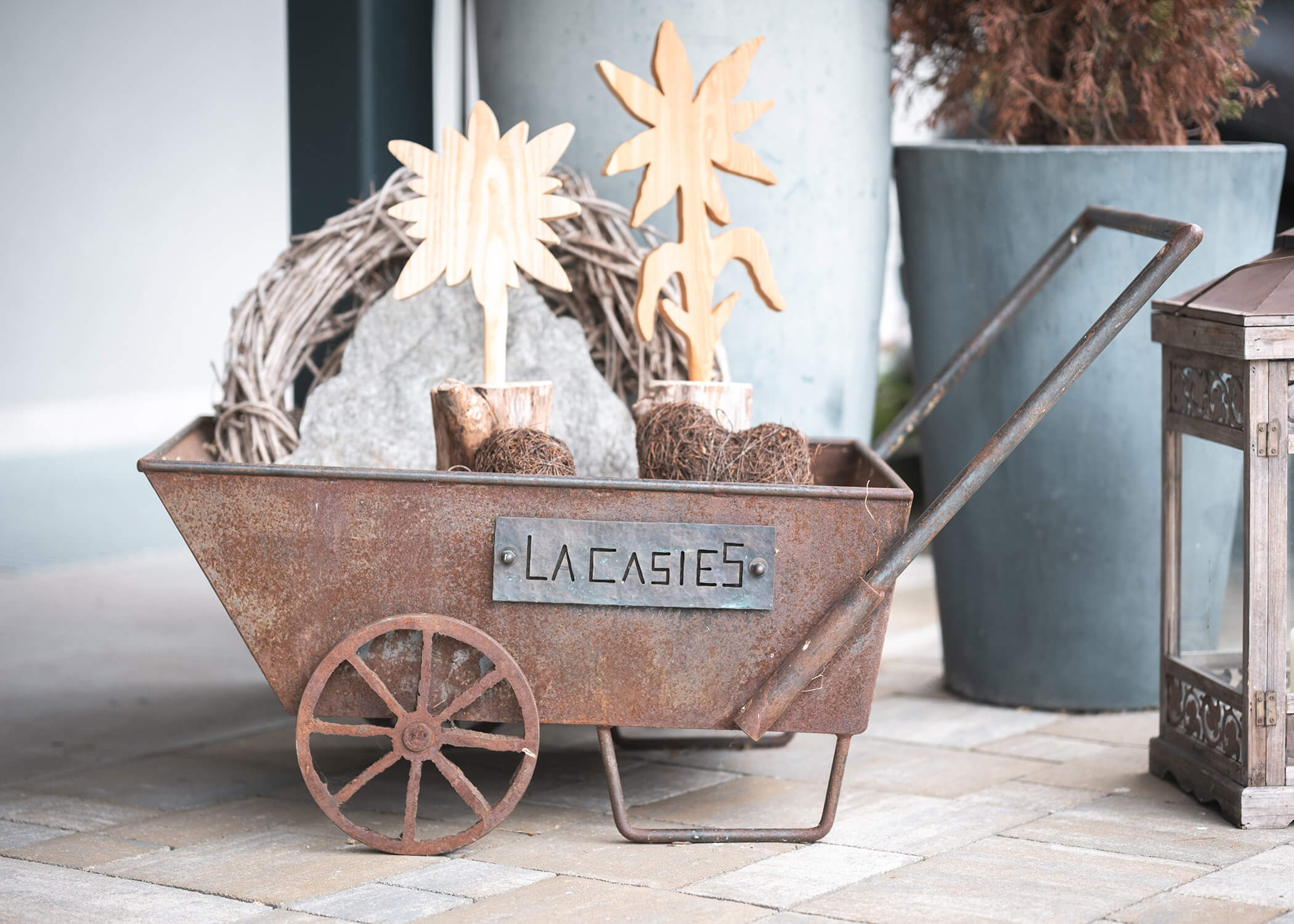 Rostiger Karren mit einem LA CASIES-Schild, auf dem sich hölzerner Pflanzenschmuck und ein Kranz befinden. - Hotel La Casies Mountain Living