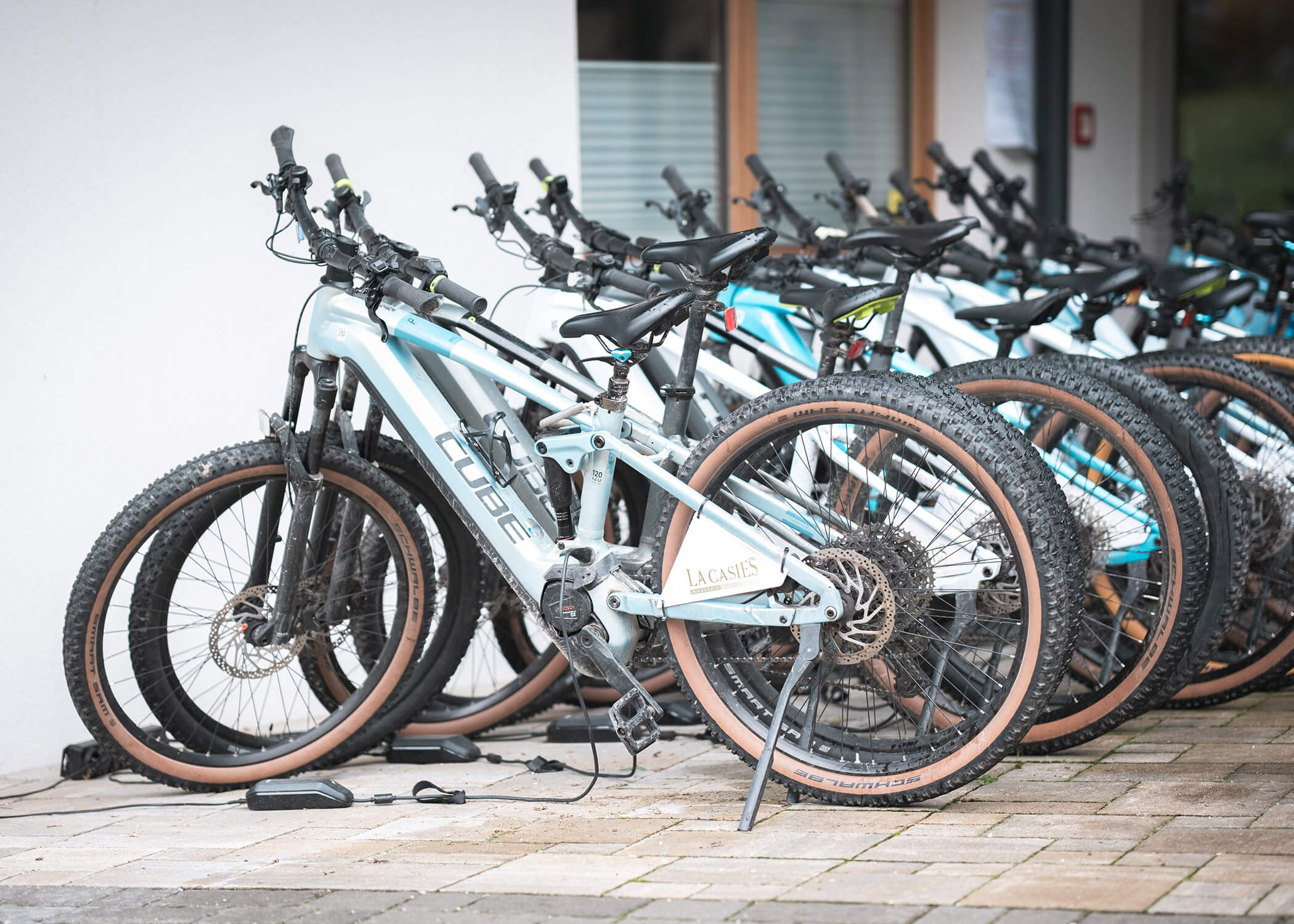 Mehrere moderne Mountainbikes sind in einer Reihe auf einer gepflasterten Fläche vor einem Gebäude abgestellt. - Hotel La Casies Mountain Living