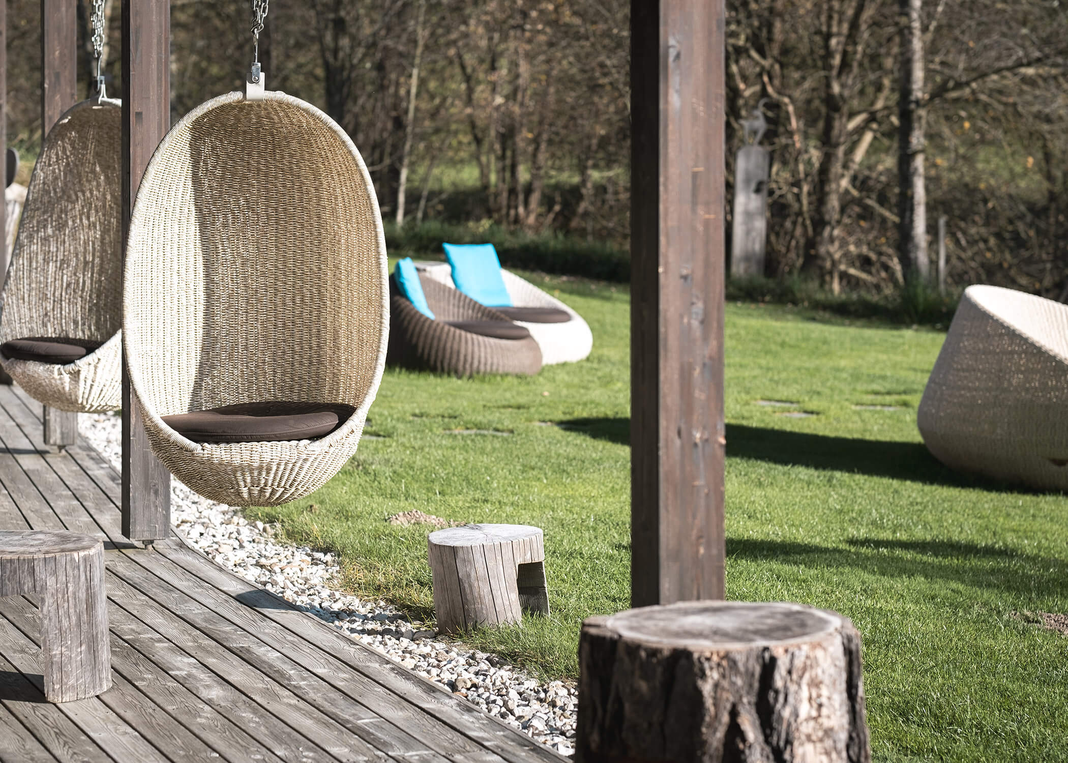 Hängesessel aus Korbgeflecht auf einer Holzterrasse mit Blick auf einen grasbewachsenen Garten mit Liegestühlen und Baumstümpfen. - Hotel La Casies Mountain Living
