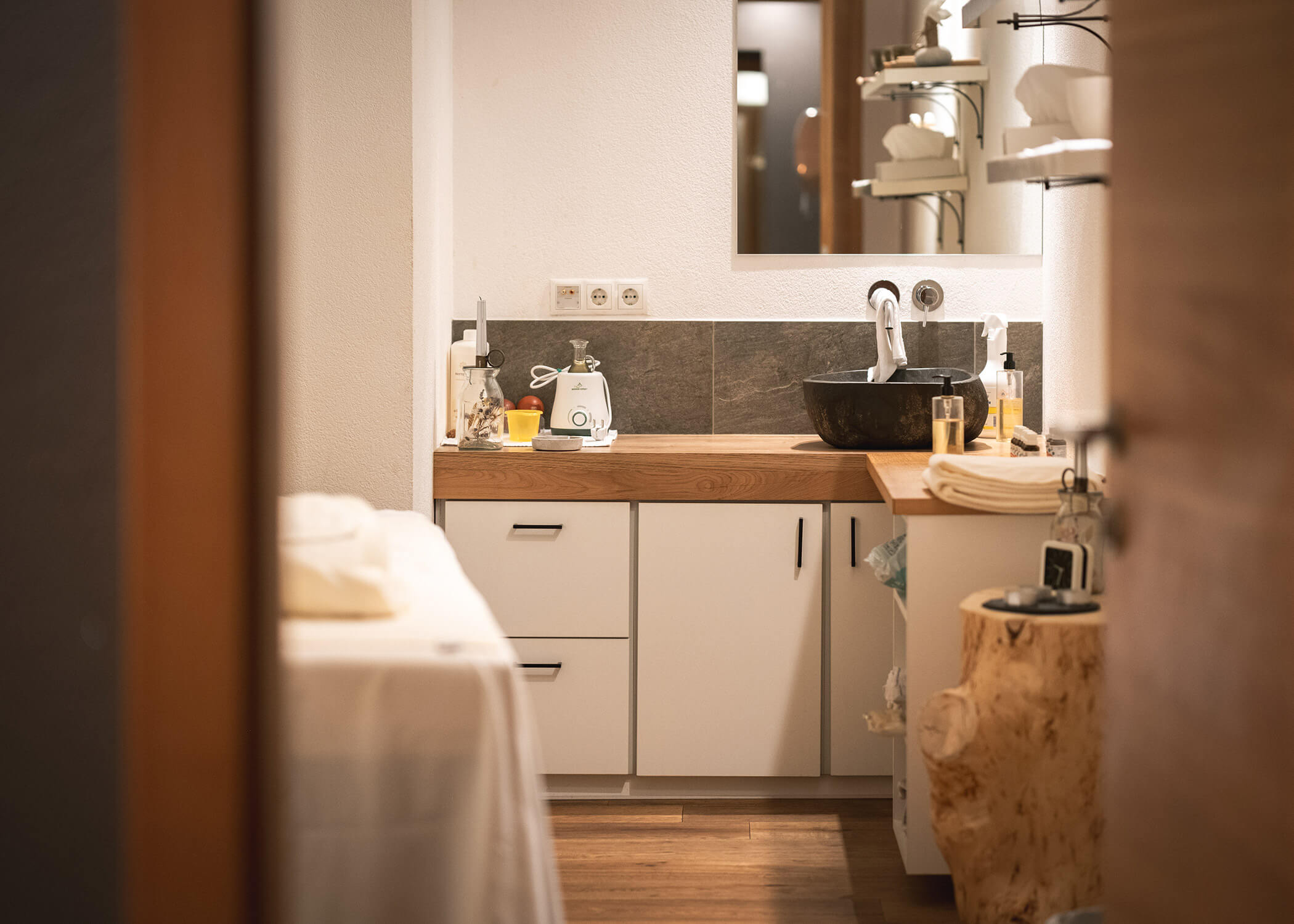 Gemütlicher Spa-Raum mit Massageliege, Holzdekor, Waschbecken, Handtüchern und Toilettenartikeln auf dem Tresen. - Hotel La Casies Mountain Living