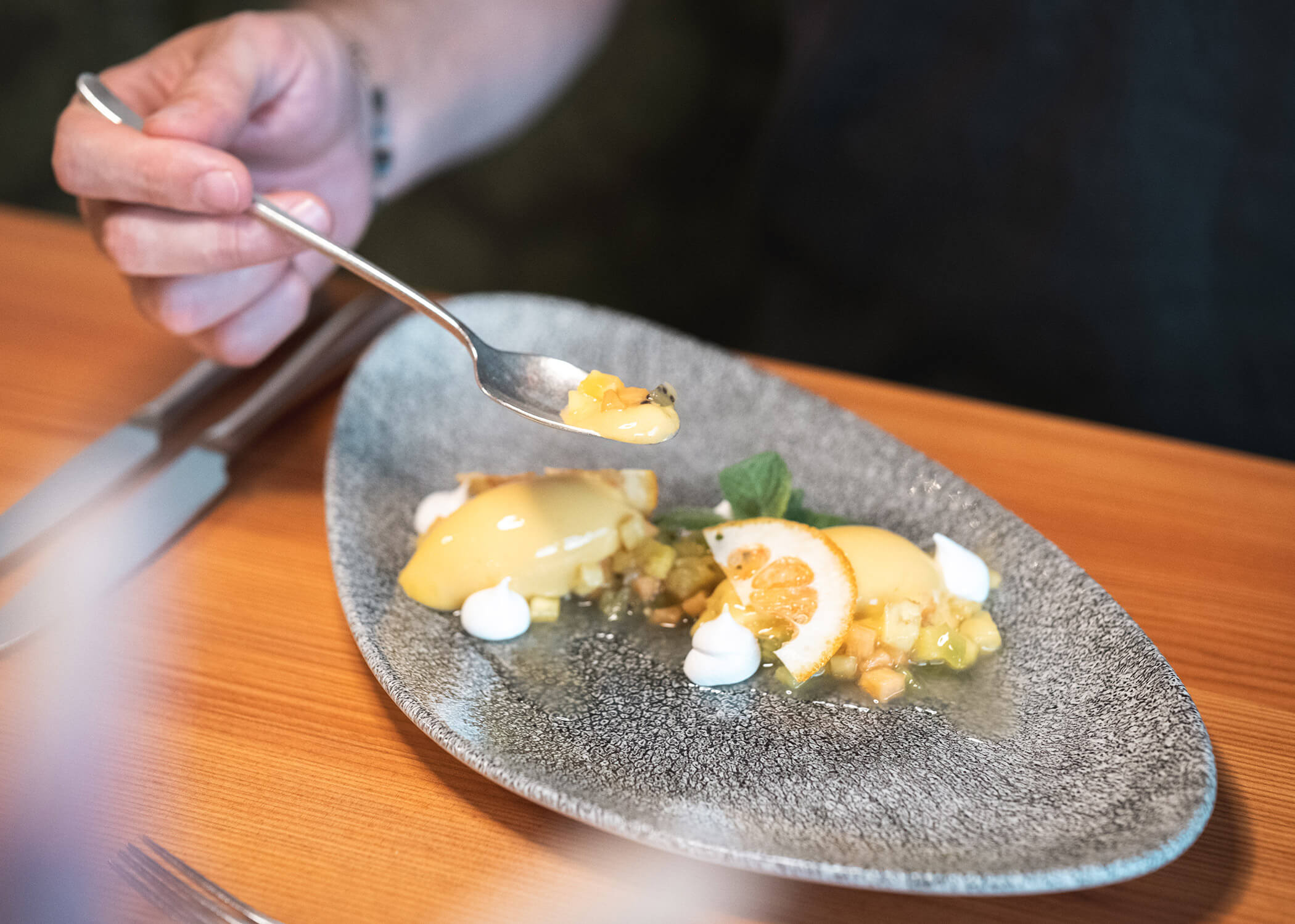 Eine Hand hält einen Löffel über ein Dessert mit Zitronenquark, Obst und Sahnetupfern auf einem grauen Teller. - Hotel La Casies Mountain Living