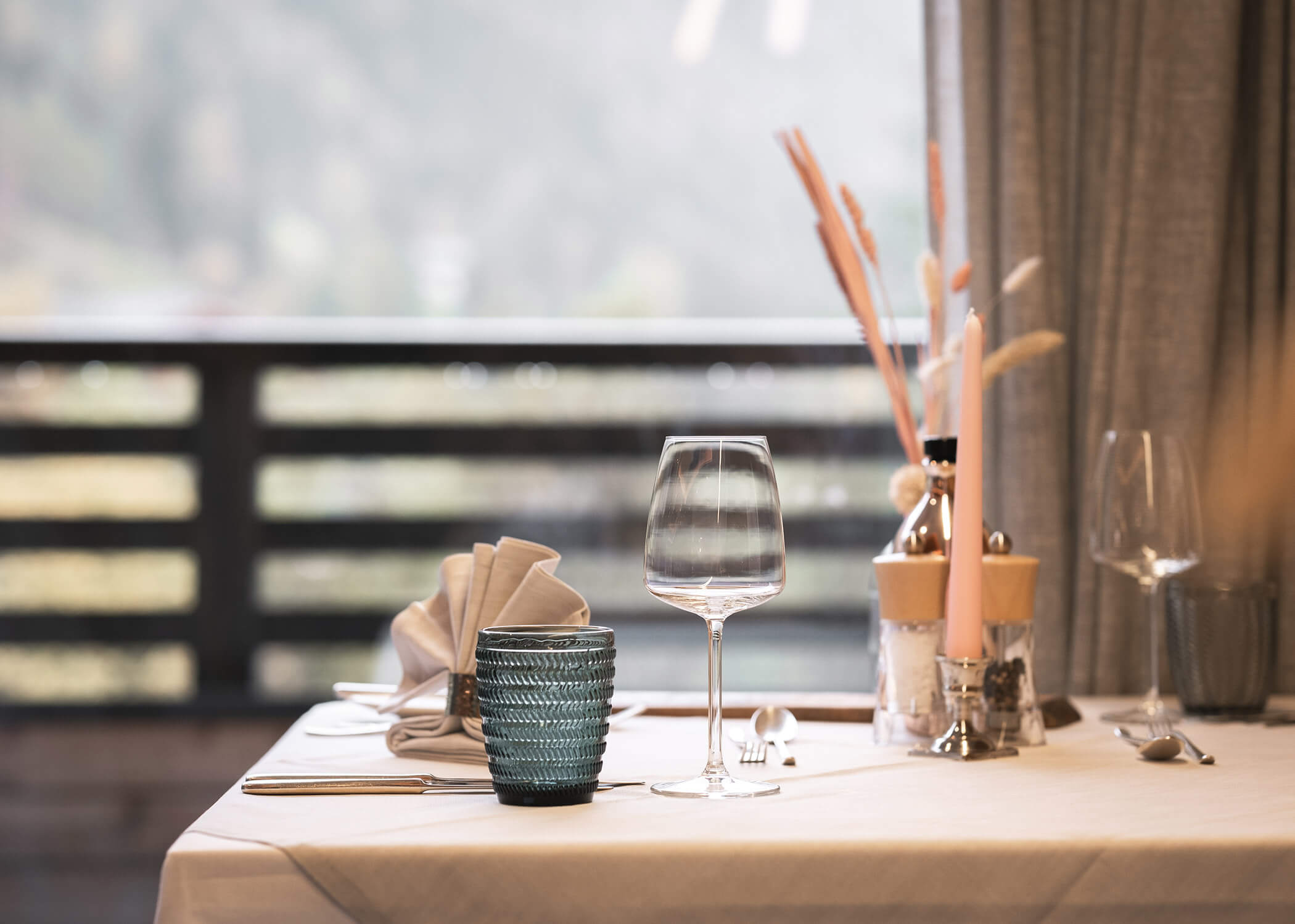 Ein ordentlich gedeckter Esstisch mit Gläsern, blauer Tasse, Besteck und Serviette an einem Fenster mit Blick auf die Landschaft. - Hotel La Casies Mountain Living