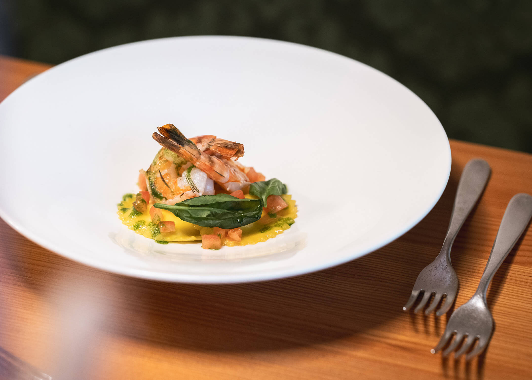 Ein weißer Teller mit Garnelen auf Nudeln und Grünzeug, garniert mit Tomatenwürfeln, daneben zwei Gabeln. - Hotel La Casies Mountain Living