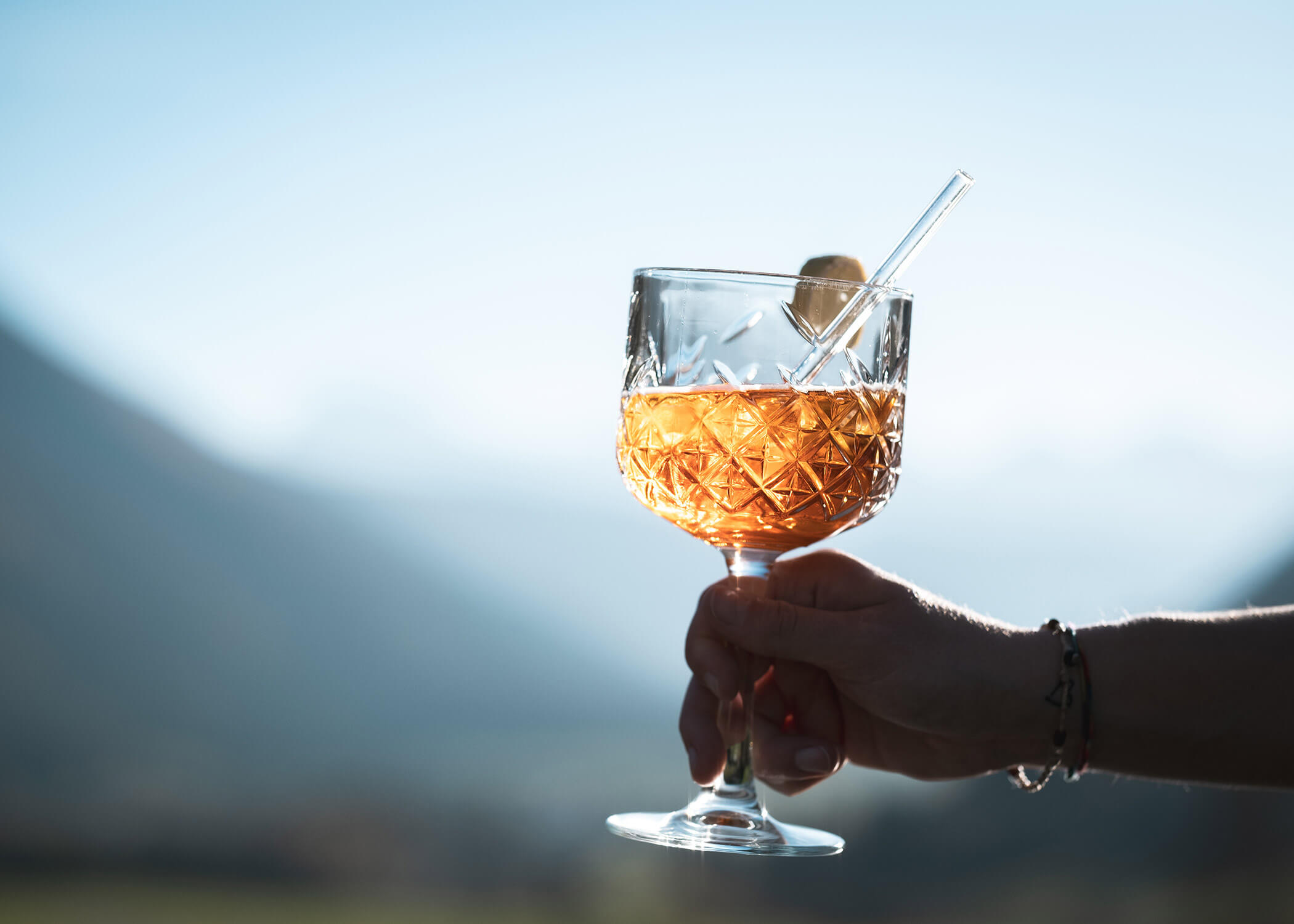 Eine Hand hält ein Kristallglas mit einem Orangencocktail und einem Strohhalm vor einem unscharfen Hintergrund im Freien. - Hotel La Casies Mountain Living