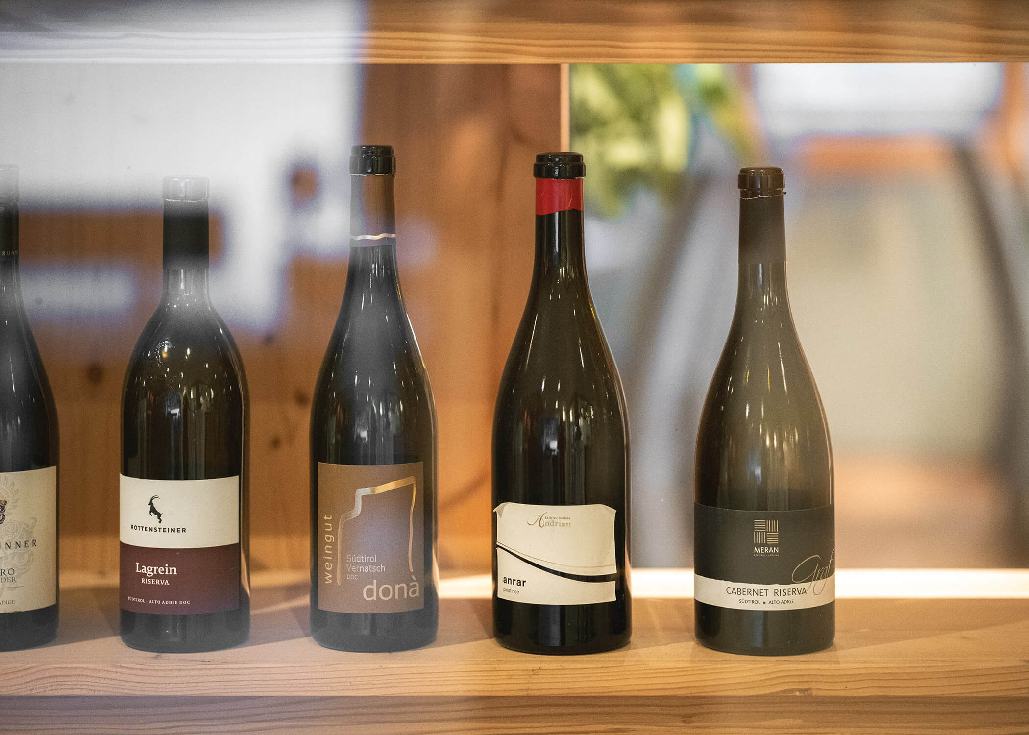 Vier Weinflaschen mit unterschiedlichen Etiketten auf einem Holzregal hinter Glas. - Hotel La Casies Mountain Living