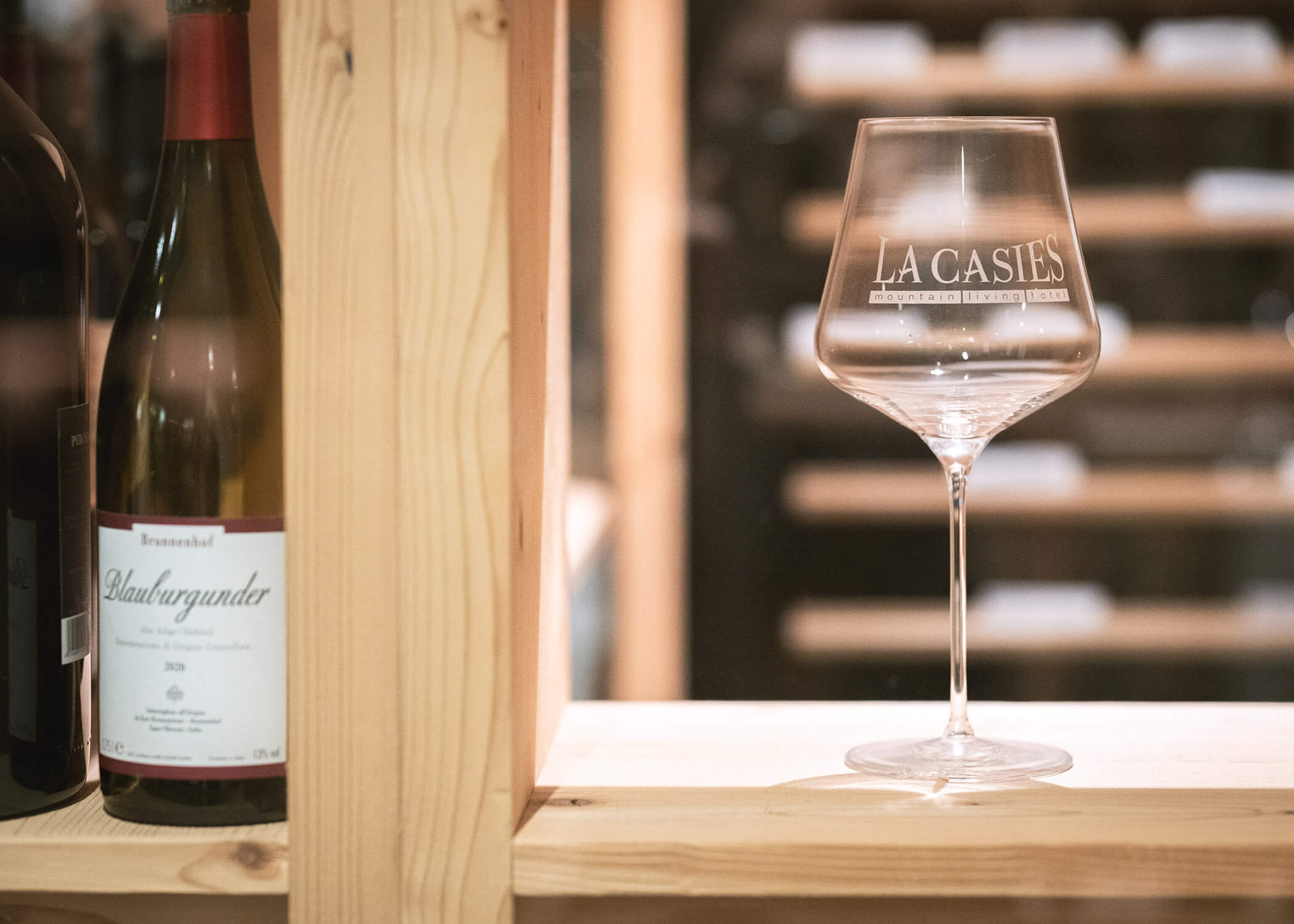Ein Weinglas mit der Aufschrift LA CASTES steht in einem Holzregal neben einer Flasche Blauer Burgunder. - Hotel La Casies Mountain Living
