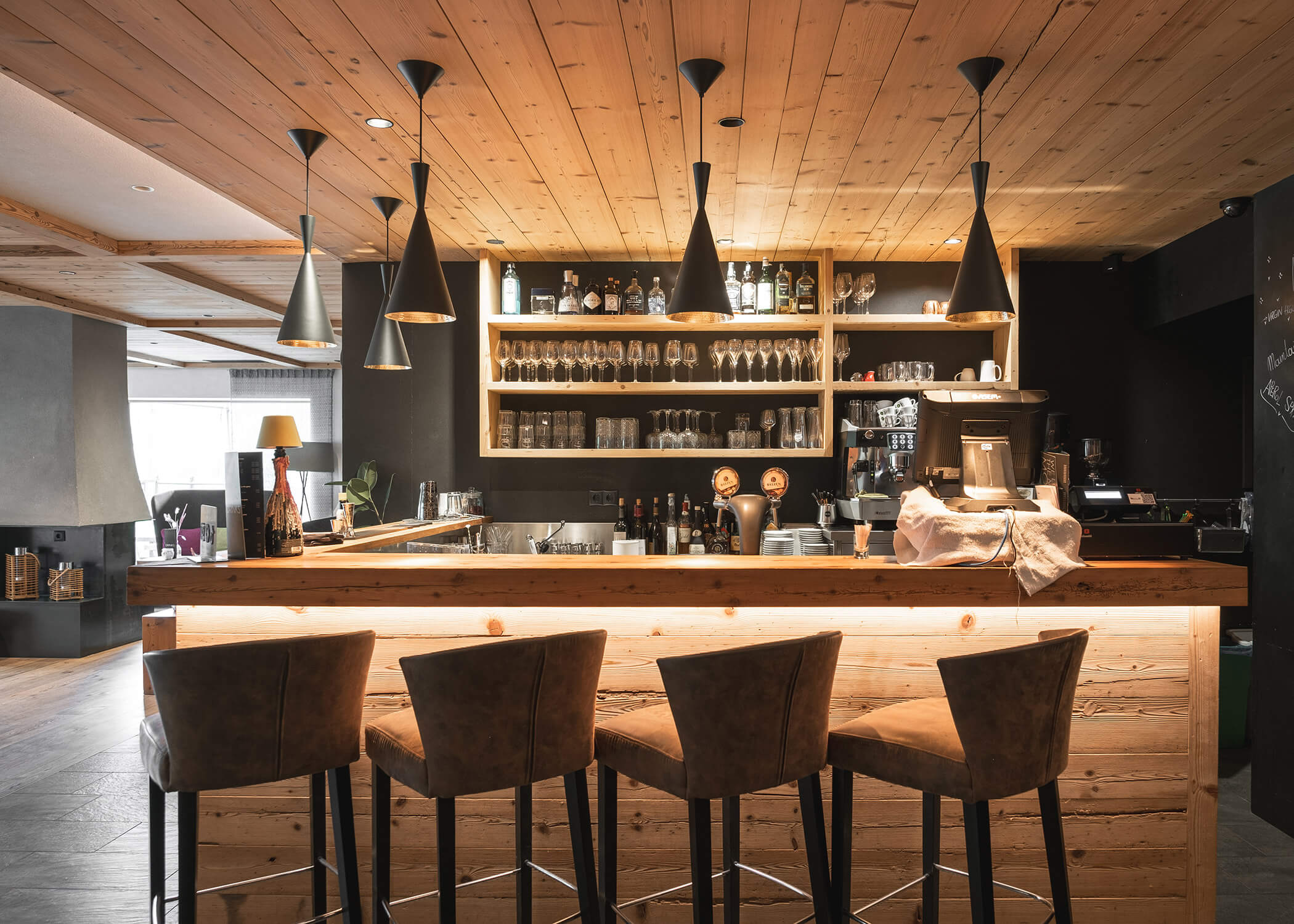 Moderne Bar mit Holzdekor, vier Barhockern, Hängelampen und Regalen mit Gläsern und Flaschen. - Hotel La Casies Mountain Living