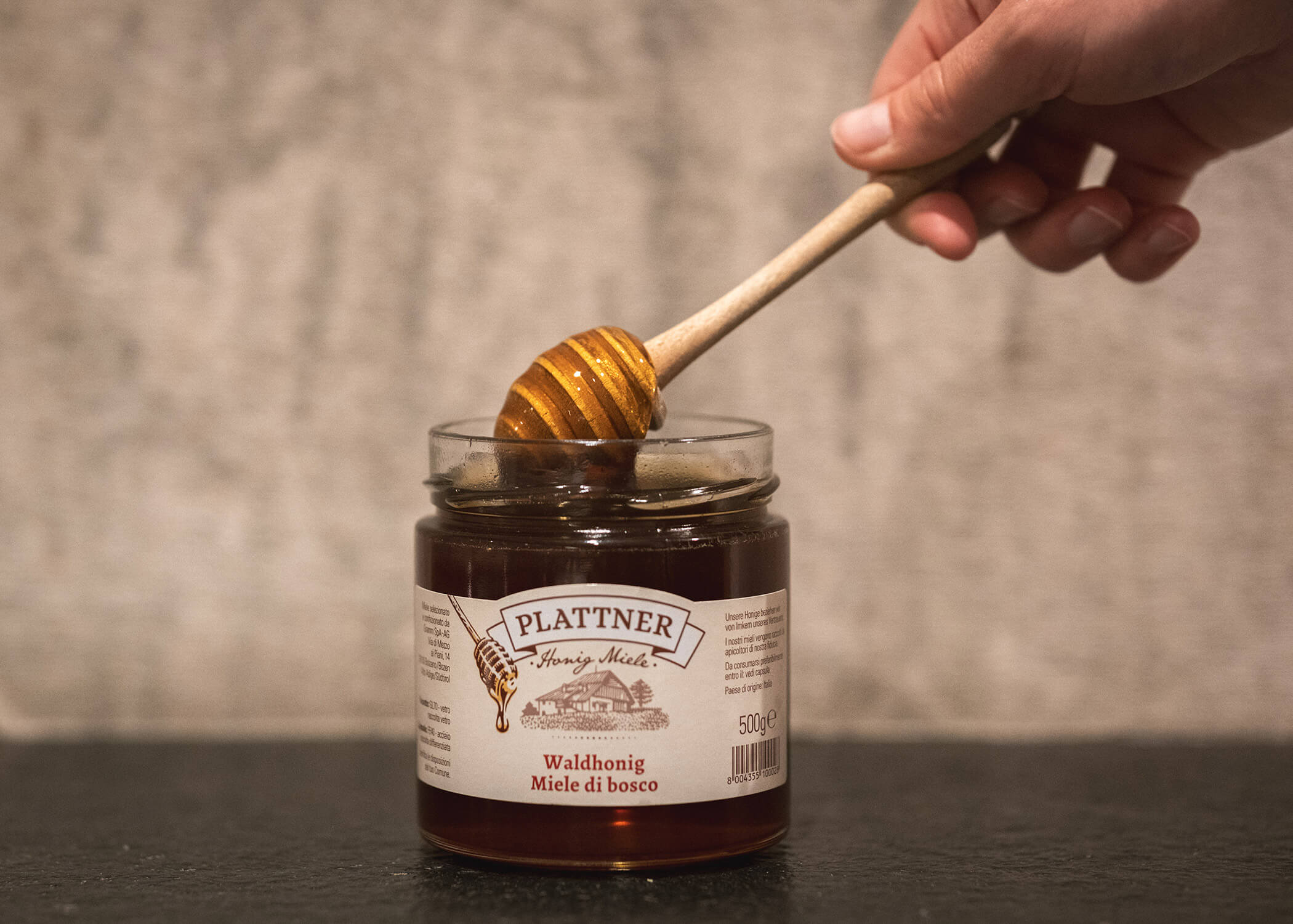 Eine Hand taucht einen Honiglöffel in ein Glas mit Plattner-Waldhonig auf einer dunklen Fläche. - Hotel La Casies Mountain Living