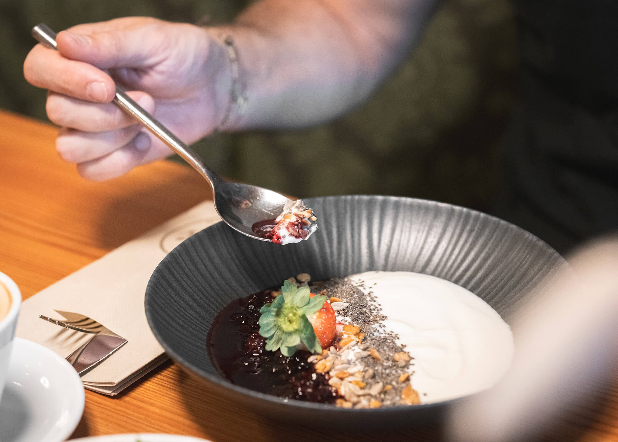 Eine Hand hält einen Löffel über eine Schüssel Joghurt mit Müsli, Samen, Beerensauce und einer Erdbeere. - Hotel La Casies Mountain Living