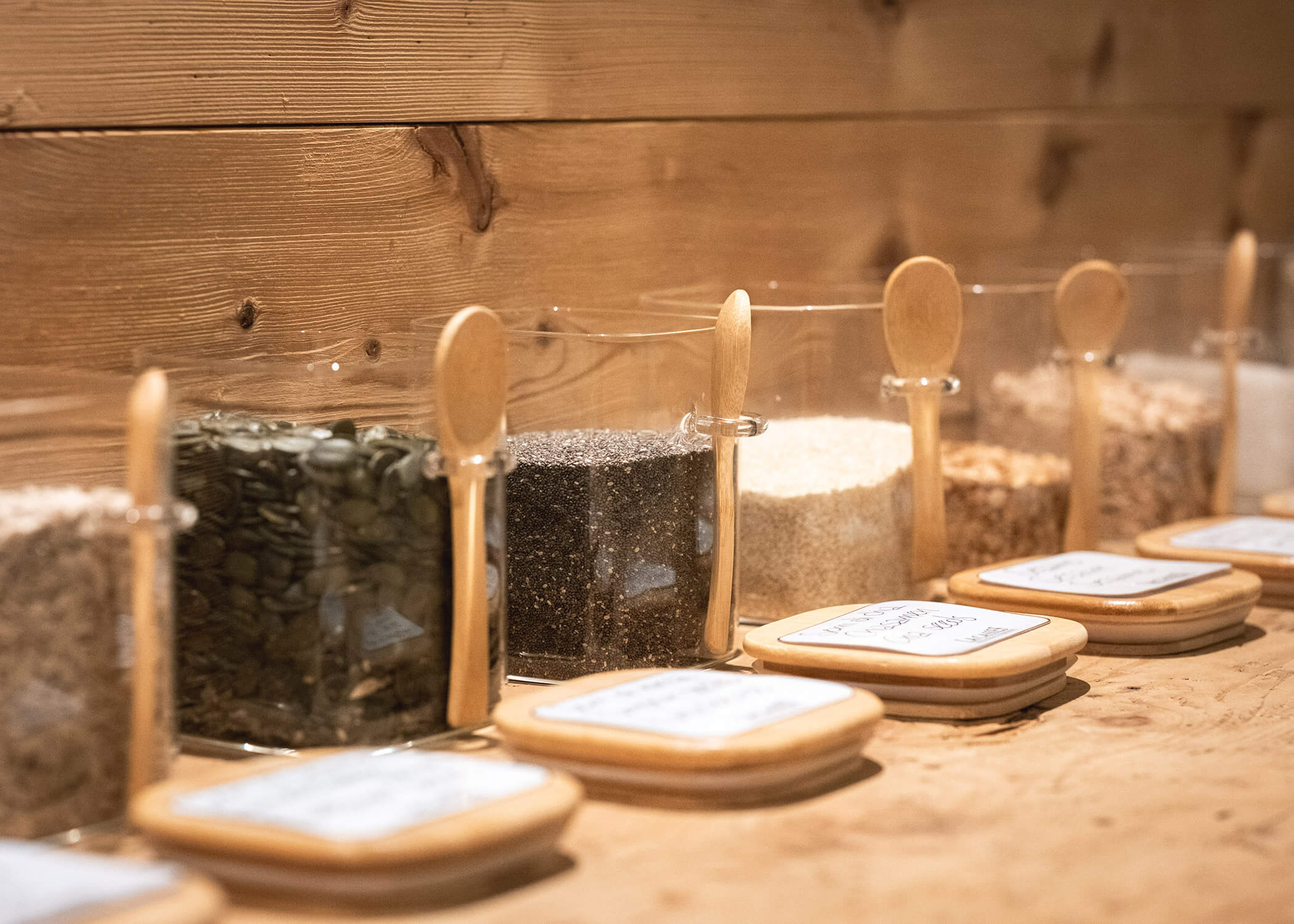 Gläser mit verschiedenen Samen und Körnern mit Holzlöffeln, aufgereiht auf einem Holztisch an einer Holzwand. - Hotel La Casies Mountain Living