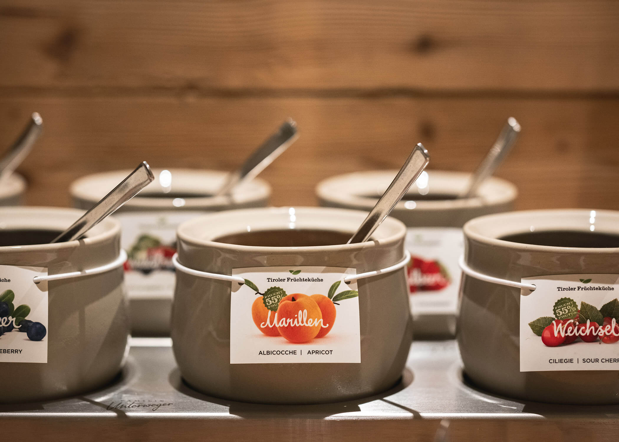 Keramische Marmeladengläser mit Löffeln, beschriftet mit Aprikose, Kirsche und Heidelbeere, auf einem Holztisch. - Hotel La Casies Mountain Living