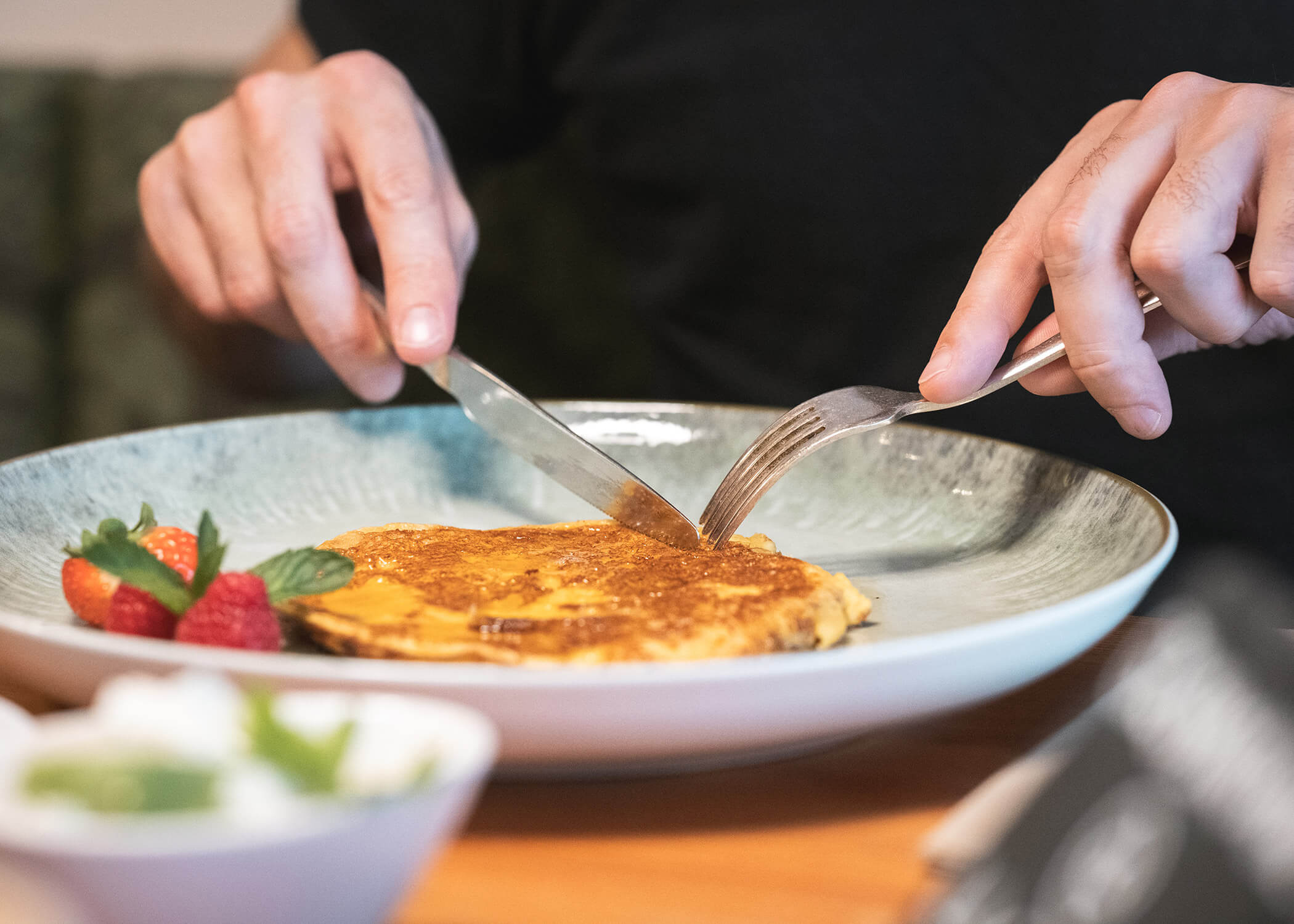 Eine Person schneidet in Pfannkuchen auf einem Teller mit Erdbeeren und Minze auf der Seite. - Hotel La Casies Mountain Living