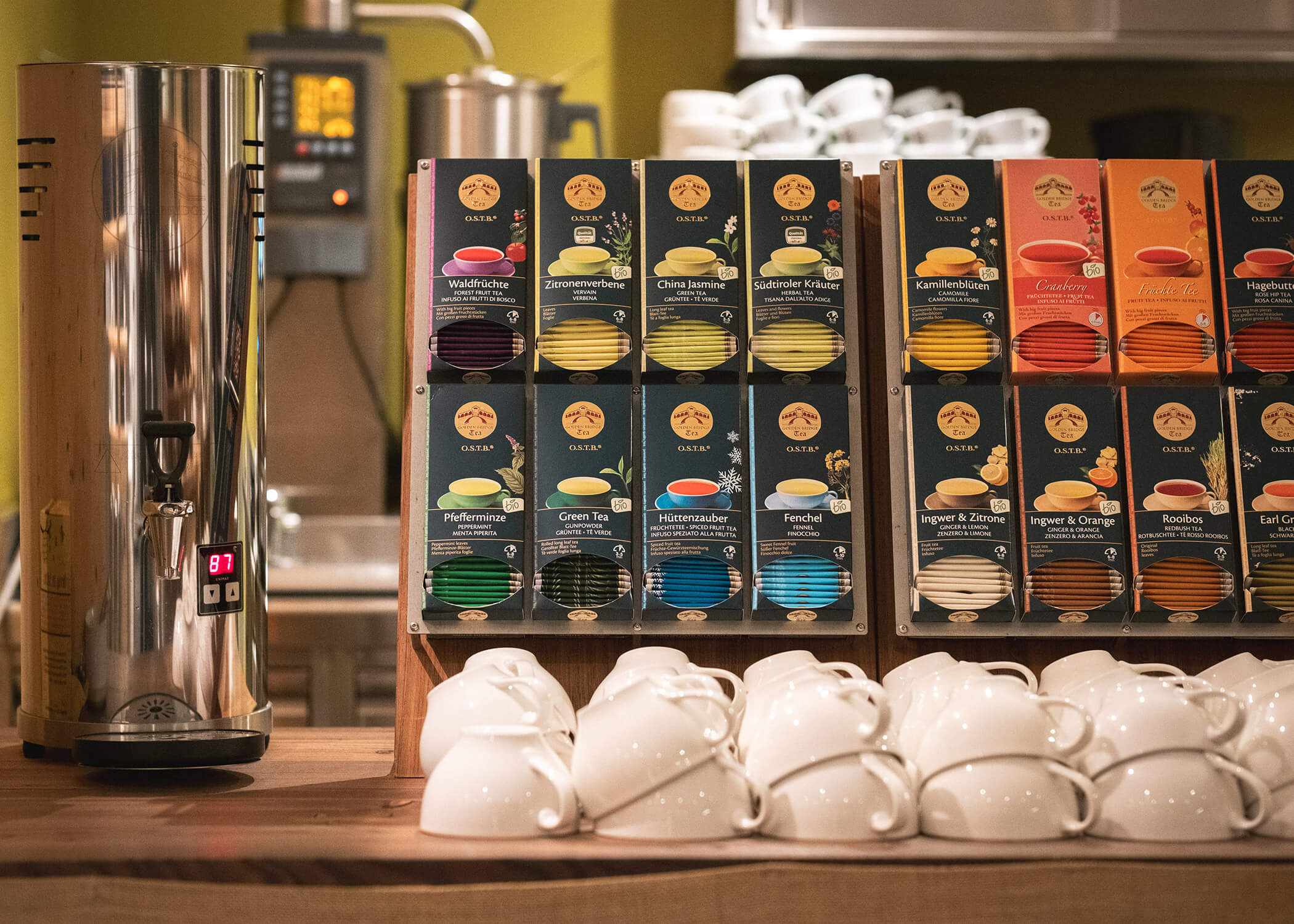 Auslage mit verschiedenen Teedosen neben gestapelten weißen Tassen und einem Heißwasserspender auf einer Holztheke. - Hotel La Casies Mountain Living