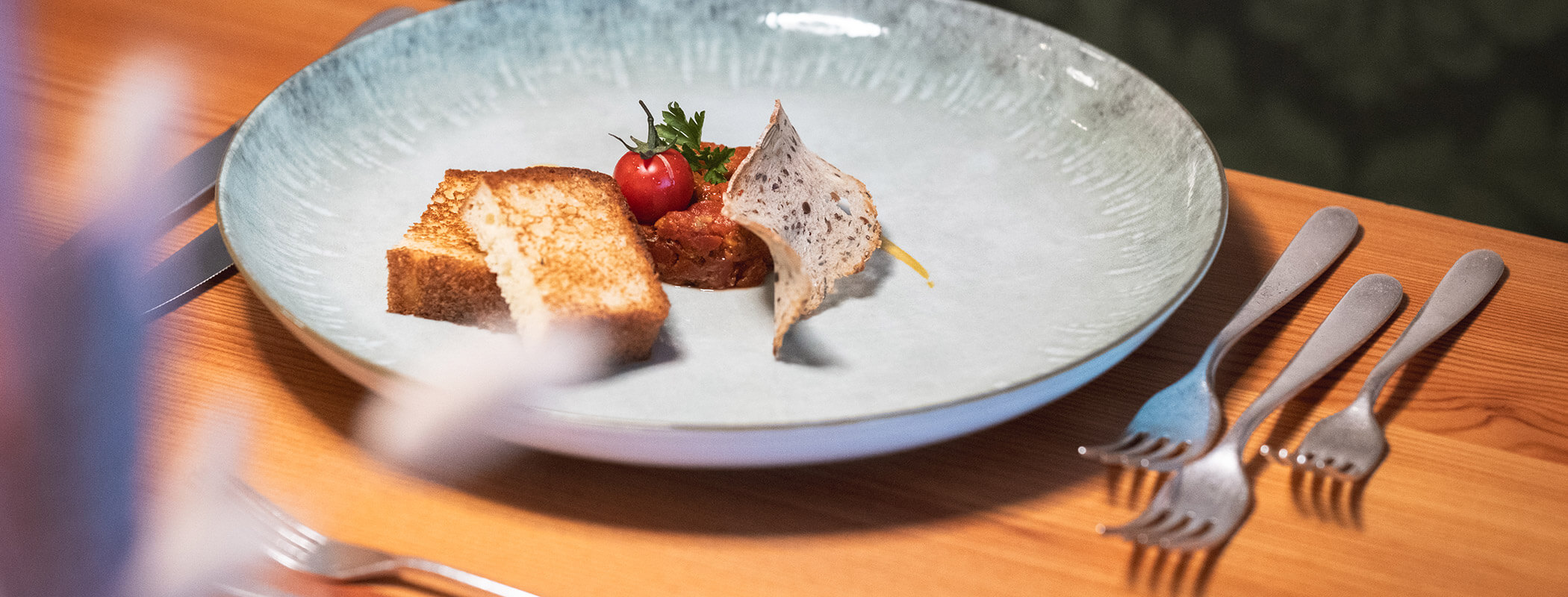 Ein Teller mit Toast, einer Tomate und einem Cracker, der auf einem Holztisch mit Gabeln und Messern steht. - Hotel La Casies Mountain Living