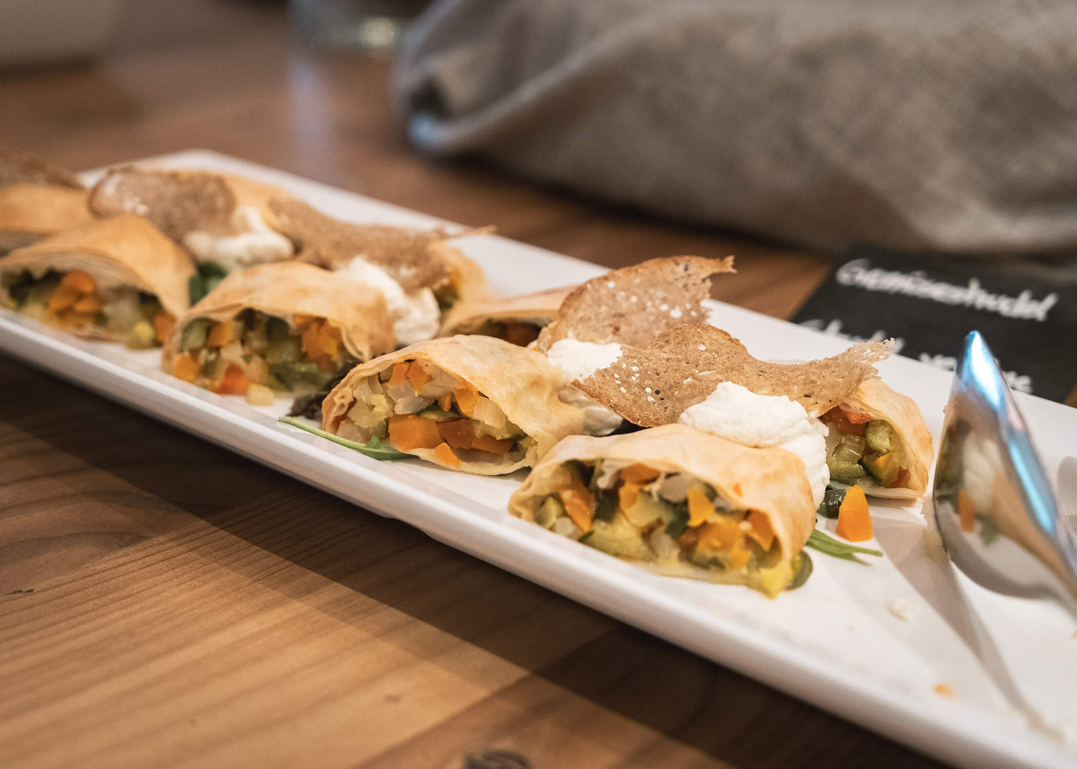 Mit Gemüse gefüllte Teigröllchen mit Sahne und knusprigen Chips, serviert auf einem weißen, rechteckigen Teller. - Hotel La Casies Mountain Living