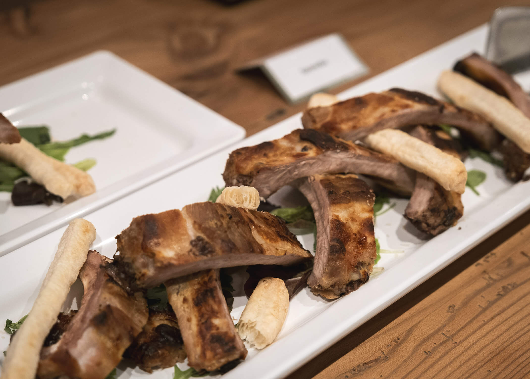 Gegrillte Rippchen und Brotstangen, serviert auf einem rechteckigen weißen Teller mit Grünzeug auf einem Holztisch. - Hotel La Casies Mountain Living