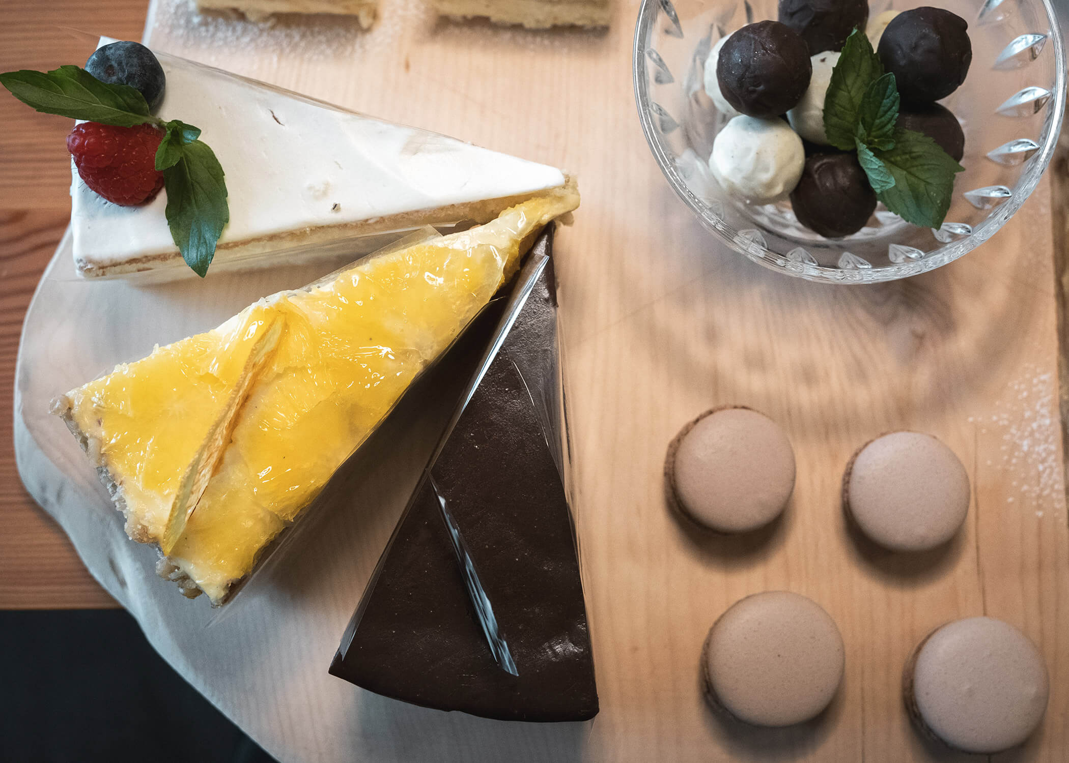 Drei Scheiben Kuchen, vier Macarons und verschiedene Pralinen in einer Glasschale auf einem Holzbrett. - Hotel La Casies Mountain Living