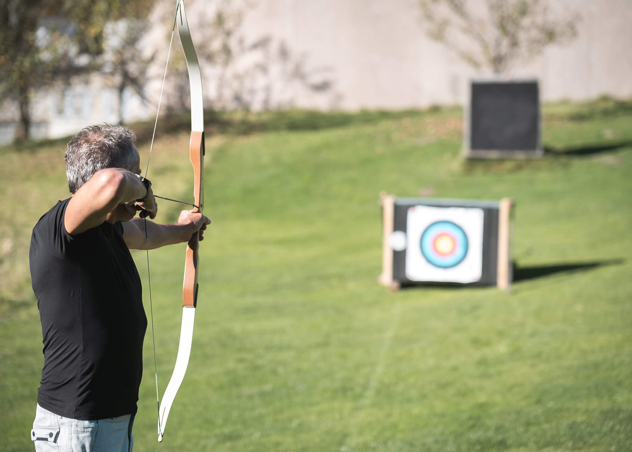 Person, die mit Pfeil und Bogen auf eine Zielscheibe auf einer Wiese zielt, während sie Bogenschießen übt. - Hotel La Casies Mountain Living