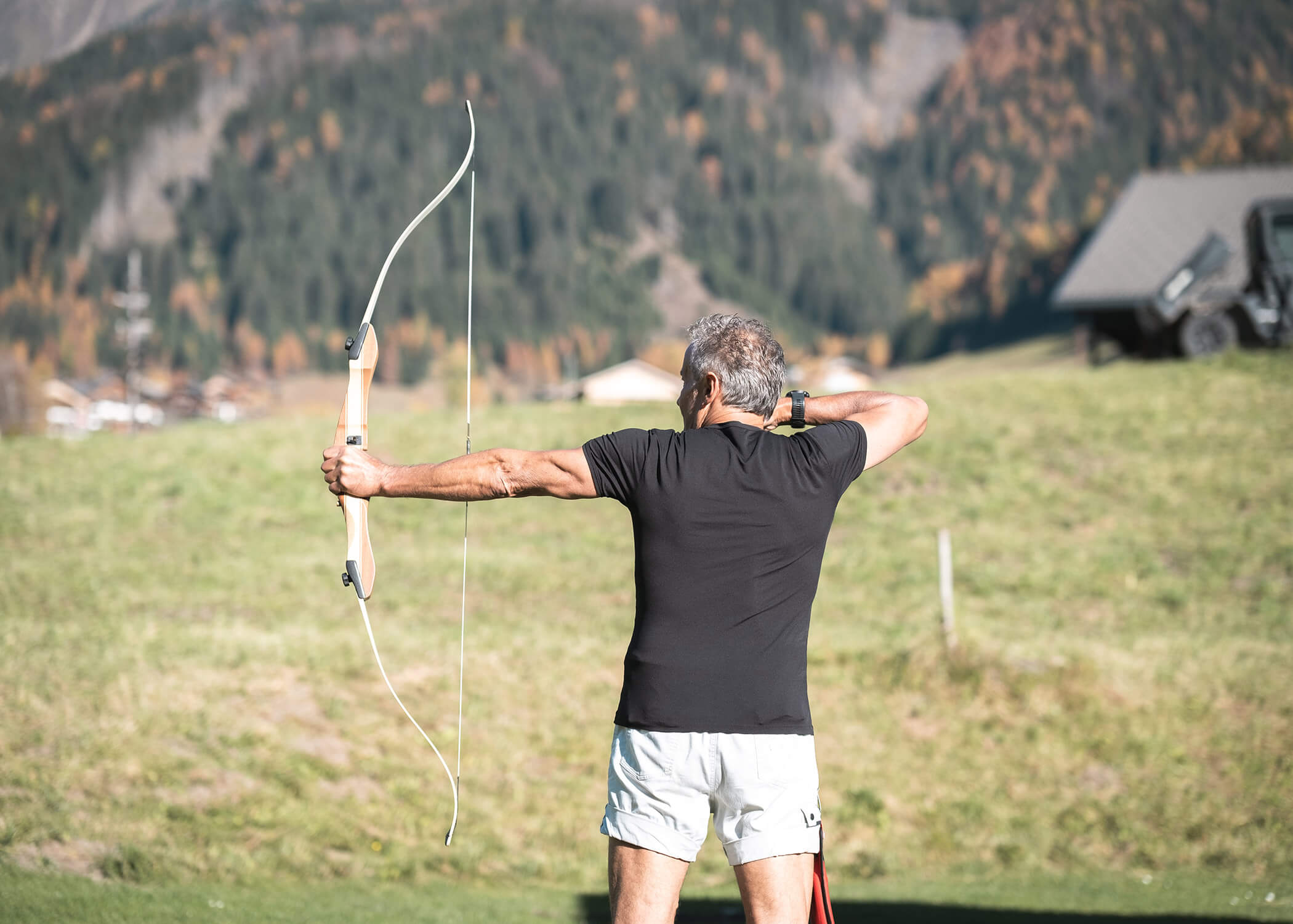 Ein Mann zielt mit einem Bogen im Freien auf einem grasbewachsenen Feld mit Bergen und Bäumen im Hintergrund. - Hotel La Casies Mountain Living