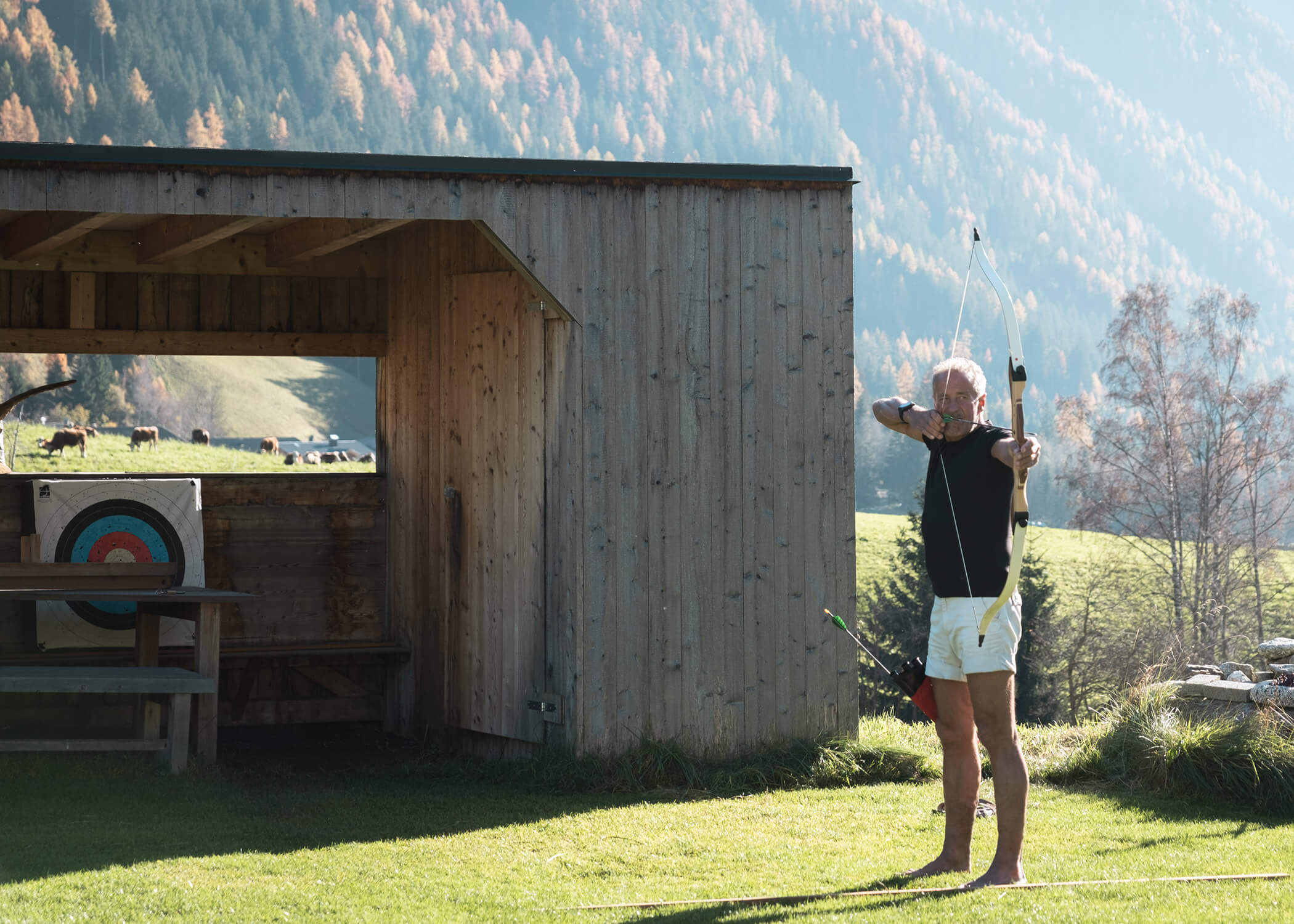 Ein barfüßiger Mann zielt mit Pfeil und Bogen auf einen Holzschuppen und eine Zielscheibe für das Bogenschießen. - Hotel La Casies Mountain Living