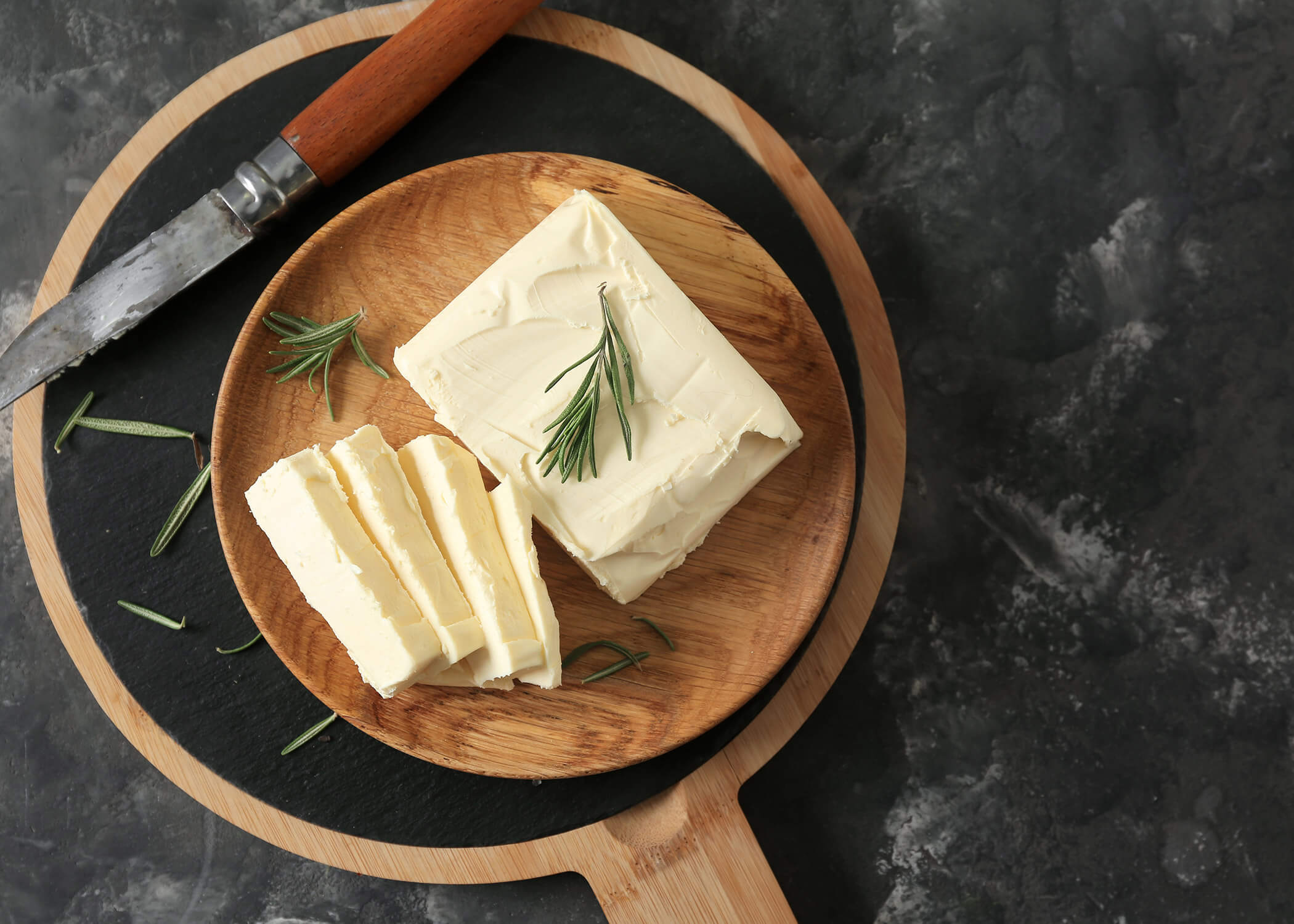 Geschnittene Butter mit Rosmarin auf einem Holzbrett, neben einem Messer auf einer dunklen Fläche. - Hotel La Casies Mountain Living