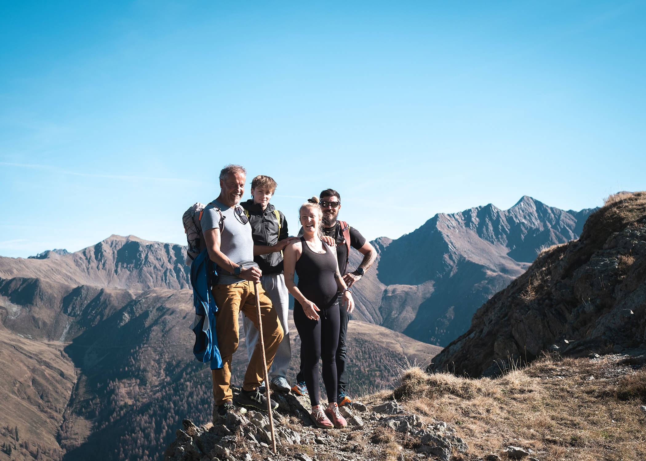 Vier Personen posieren auf einem felsigen Bergpfad mit weit entfernten Gipfeln und einem klaren blauen Himmel im Hintergrund. - Hotel La Casies Mountain Living