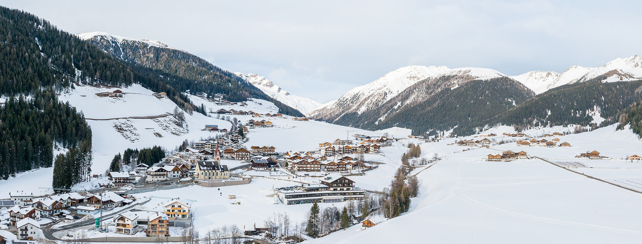 Verschneites Alpendorf in einem Tal mit Bergen und Kiefernwäldern im Hintergrund. - Hotel La Casies Mountain Living