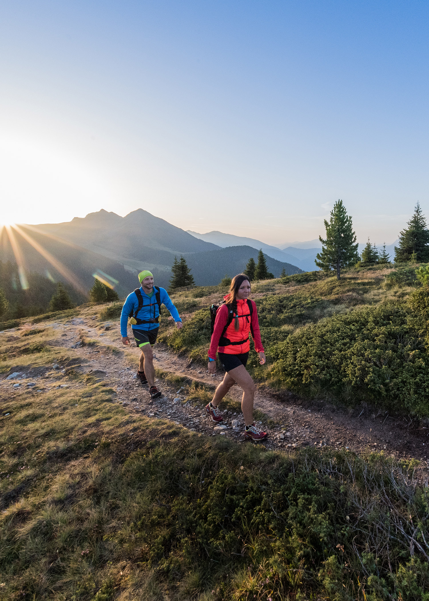 Zwei Personen, die bei Sonnenaufgang mit Rucksäcken auf einem Bergpfad wandern, umgeben von Bäumen und malerischen Aussichten. - Hotel La Casies Mountain Living