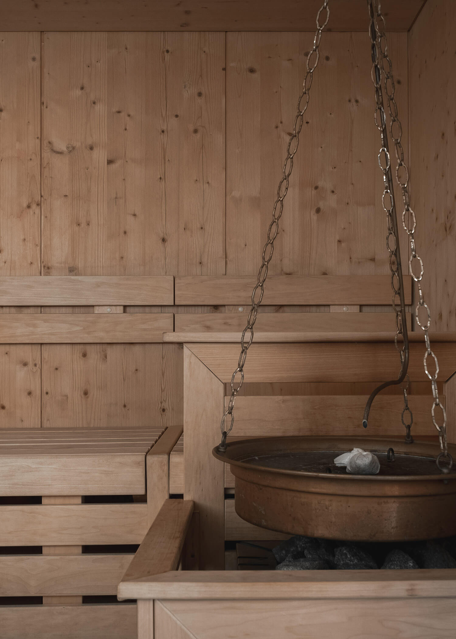 Innenraum der Holzsauna mit abgestuften Bänken und einem Metallbecken, das über den Saunasteinen hängt. - Hotel La Casies Mountain Living