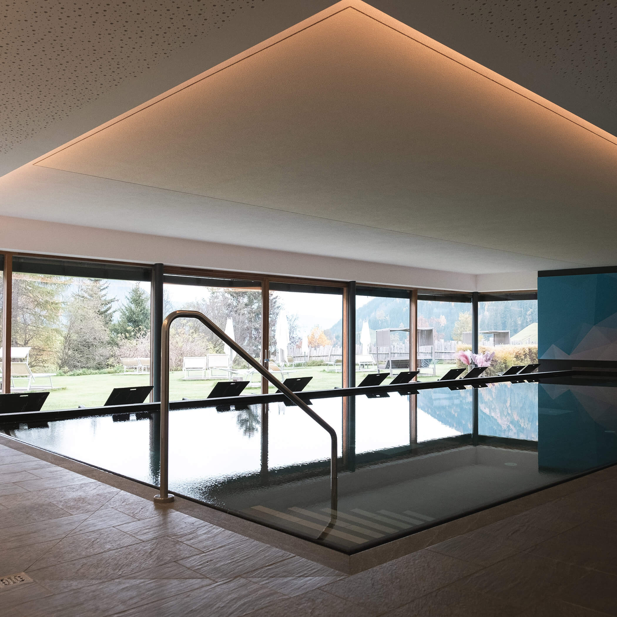 Innenpool mit Liegestühlen an großen Fenstern mit Blick auf einen grünen Garten und Bäume im Tageslicht. - Hotel La Casies Mountain Living