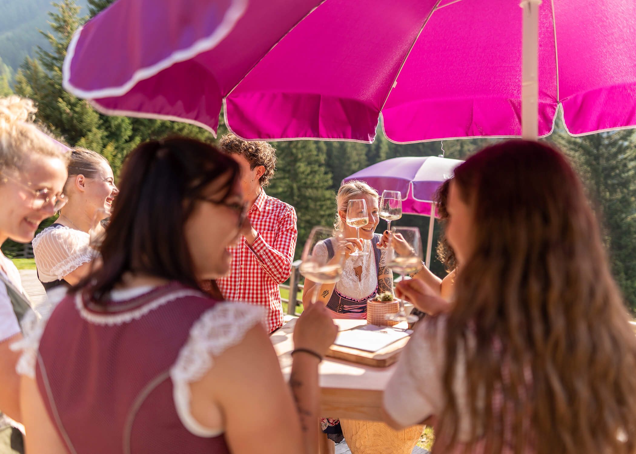 Eine Gruppe von Menschen stößt an einem sonnigen Tag unter rosa Regenschirmen mit Getränken an. - Hotel La Casies Mountain Living