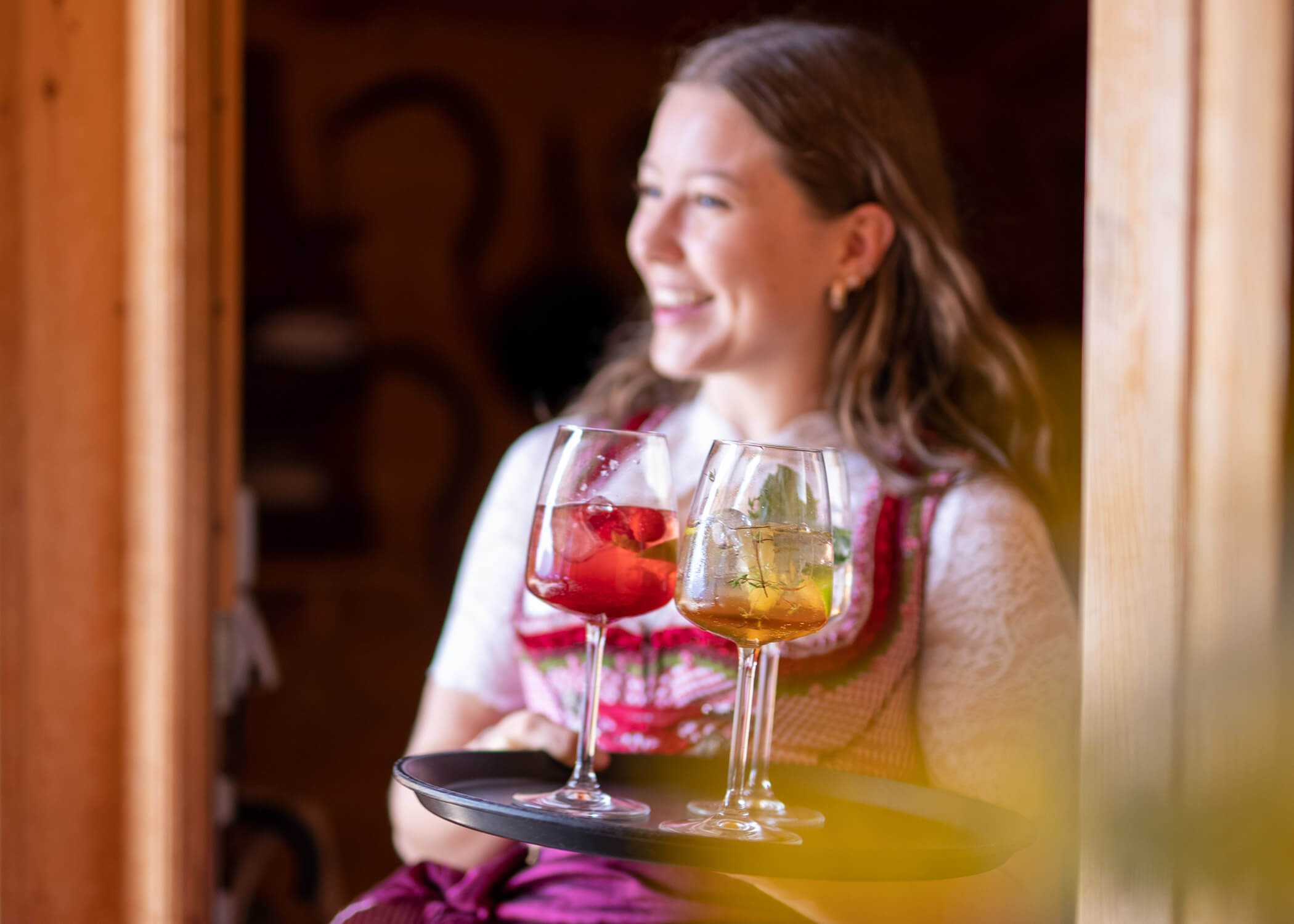 Eine lächelnde Frau in traditioneller Kleidung trägt ein Tablett mit drei bunten Getränken in Weingläsern. - Hotel La Casies Mountain Living