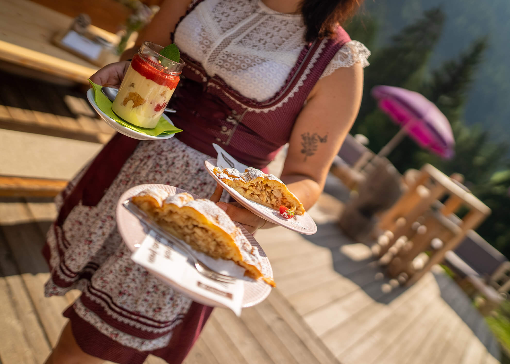 Eine Frau in traditioneller Kleidung hält an einem sonnigen Tag im Freien einen Dessertteller und ein Glas mit geschichtetem Parfait. - Hotel La Casies Mountain Living