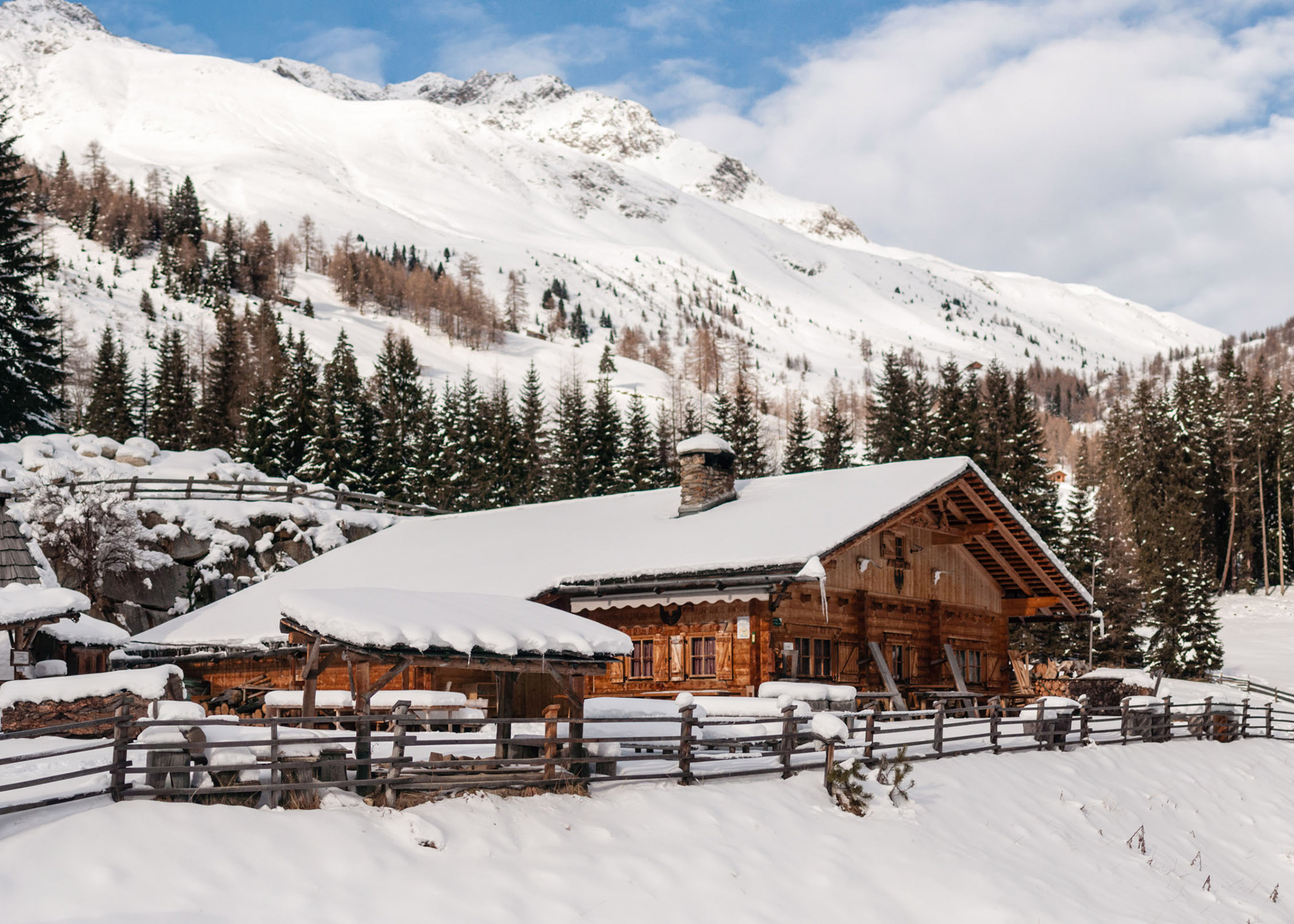 Eine Holzhütte mit schneebedecktem Dach liegt zwischen Kiefern und schneebedeckten Bergen unter einem teilweise bewölkten Himmel. - Hotel La Casies Mountain Living