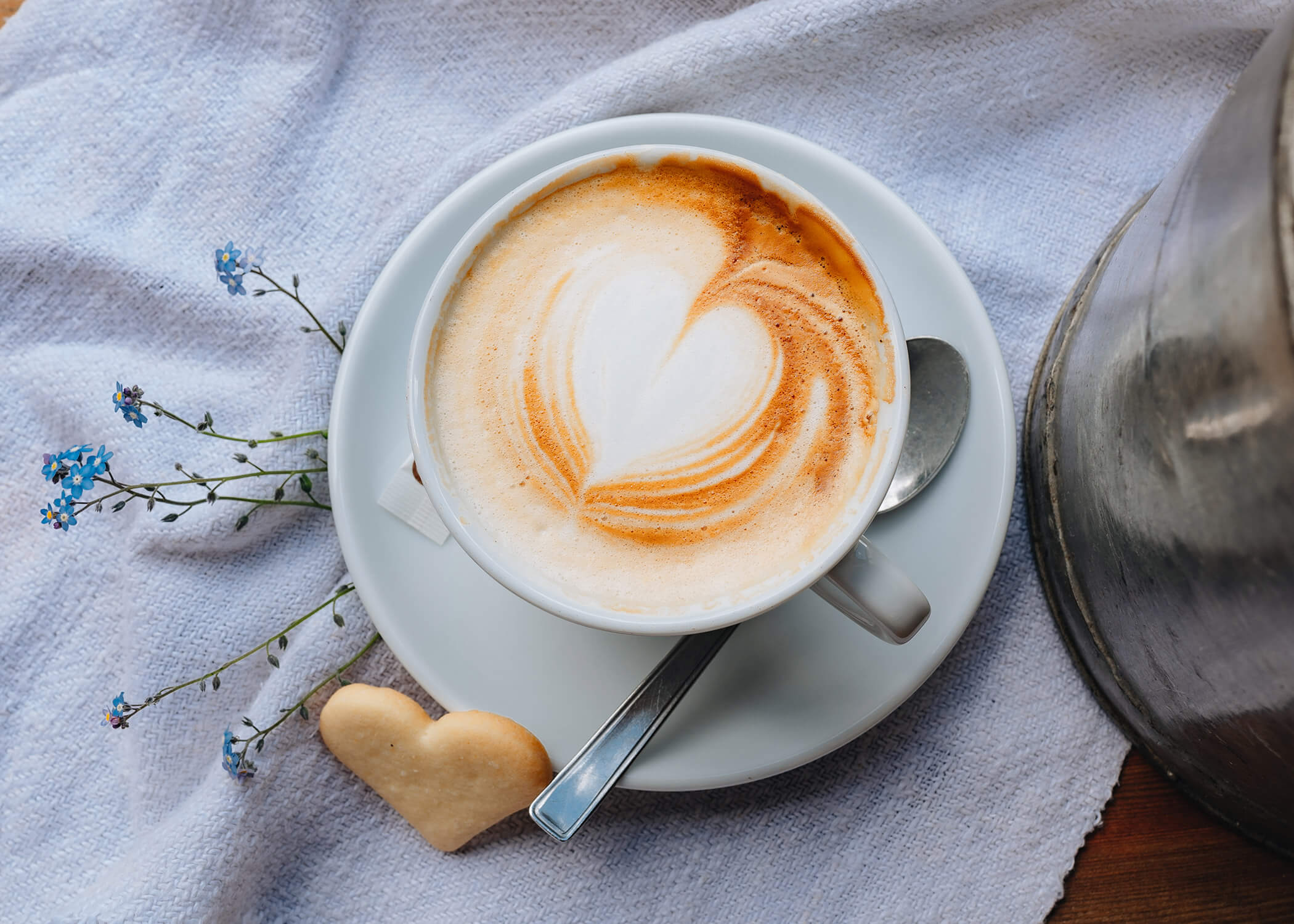 Eine Tasse Milchkaffee mit herzförmigem Schaum, ein Herzkeks und ein Löffel auf einer weißen Untertasse, auf einem Tuch. - Hotel La Casies Mountain Living