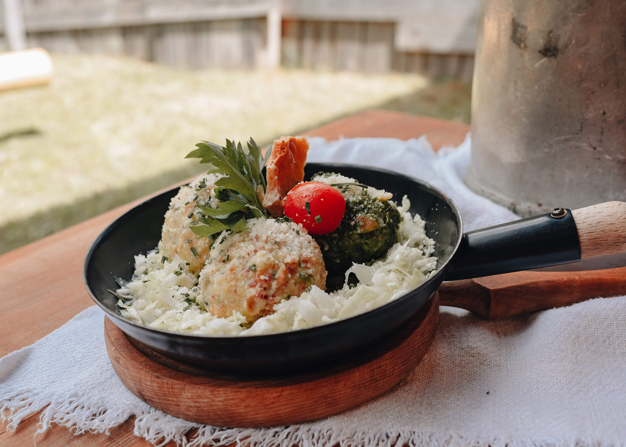 Eine schwarze Pfanne mit käsigen Gemüsebällchen, geraspeltem Kohl, Petersilie und einer Kirschtomate auf einem Holzbrett. - Hotel La Casies Mountain Living